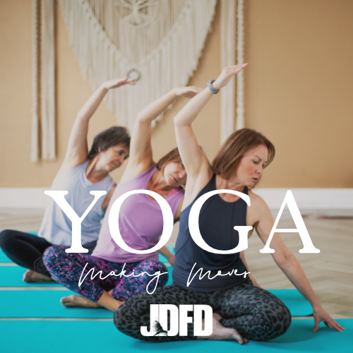 A group of three women practicing yoga on mats, stretching their arms and bodies in an indoor studio setting."