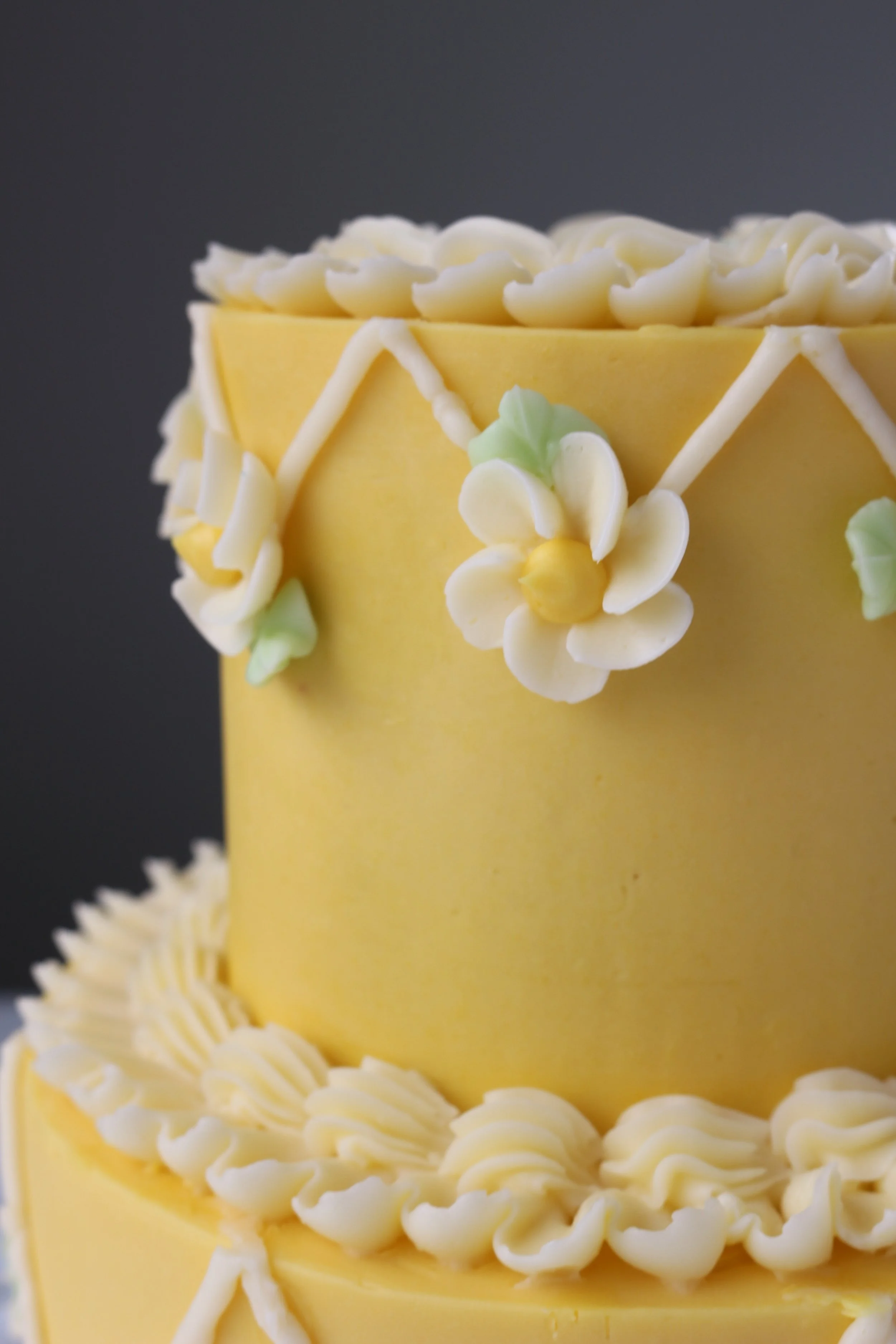 Made by Cakes by Caroline. Close-up of wo-tiered yellow cake with geometric, floral, ruffle, and shell designs.