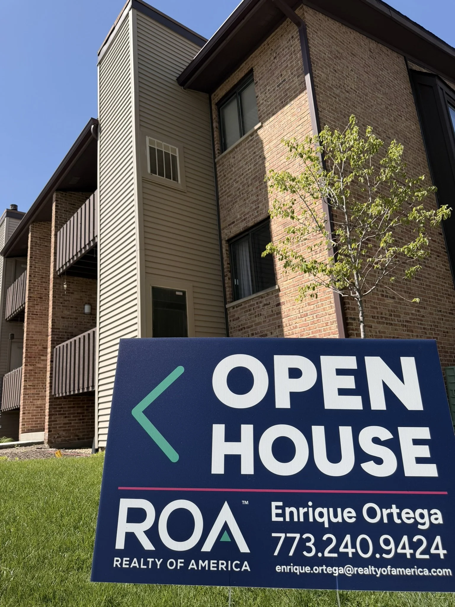 Real estate sign in front of a building with brick and siding exterior, advertising a house for sale by Enrique Ortega of Realty of America.