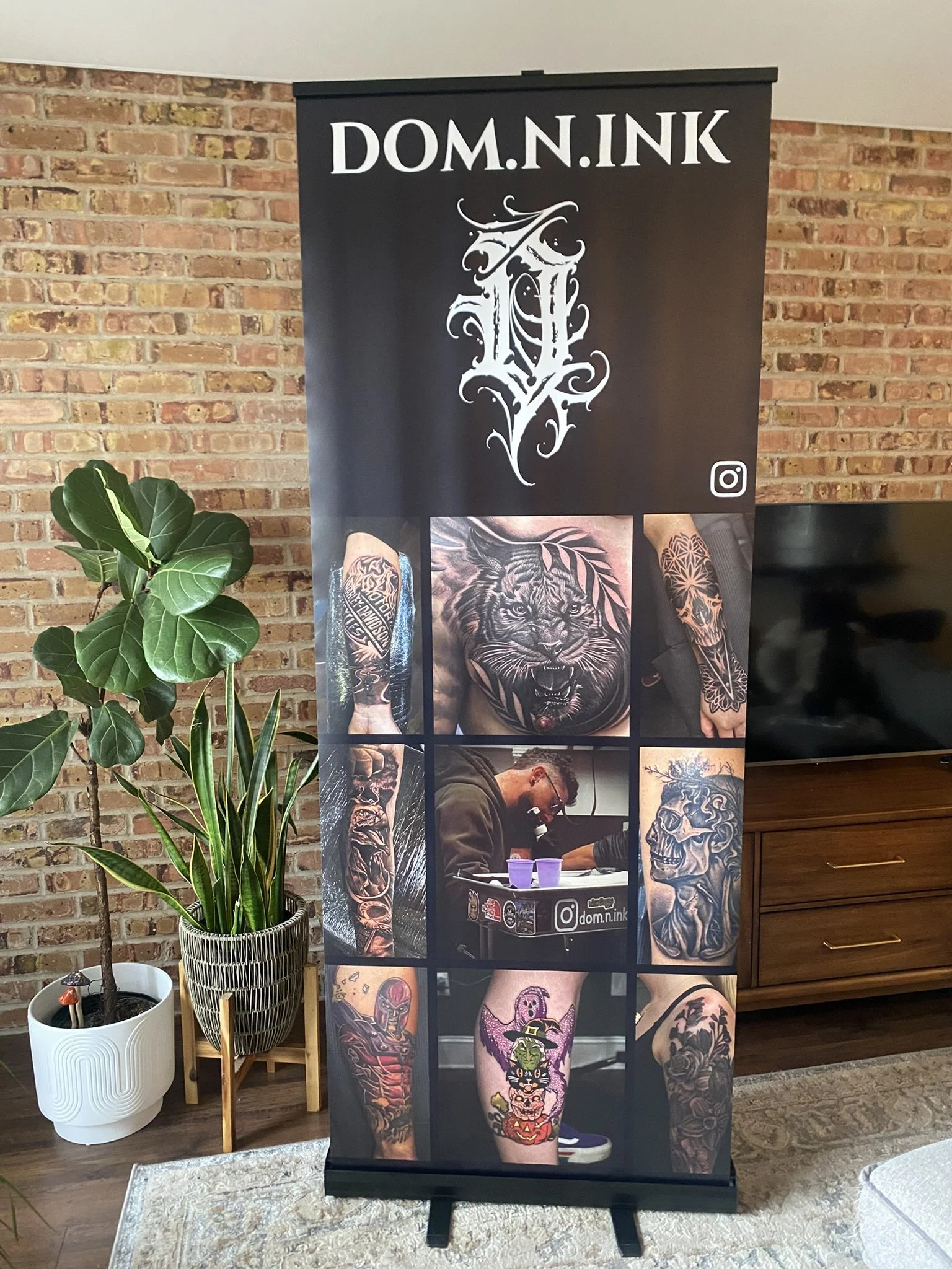 A tattoo artist is working at a tattoo booth with a banner displaying the Instagram handle 'dom.n.ink'. The banner features a collection of tattooed arms and legs, showcasing various tattoo styles, including a large lion's face, colorful cartoon characters, and intricate black and gray designs. The setting has a brick wall background with indoor plants, a wooden side table, and a TV.