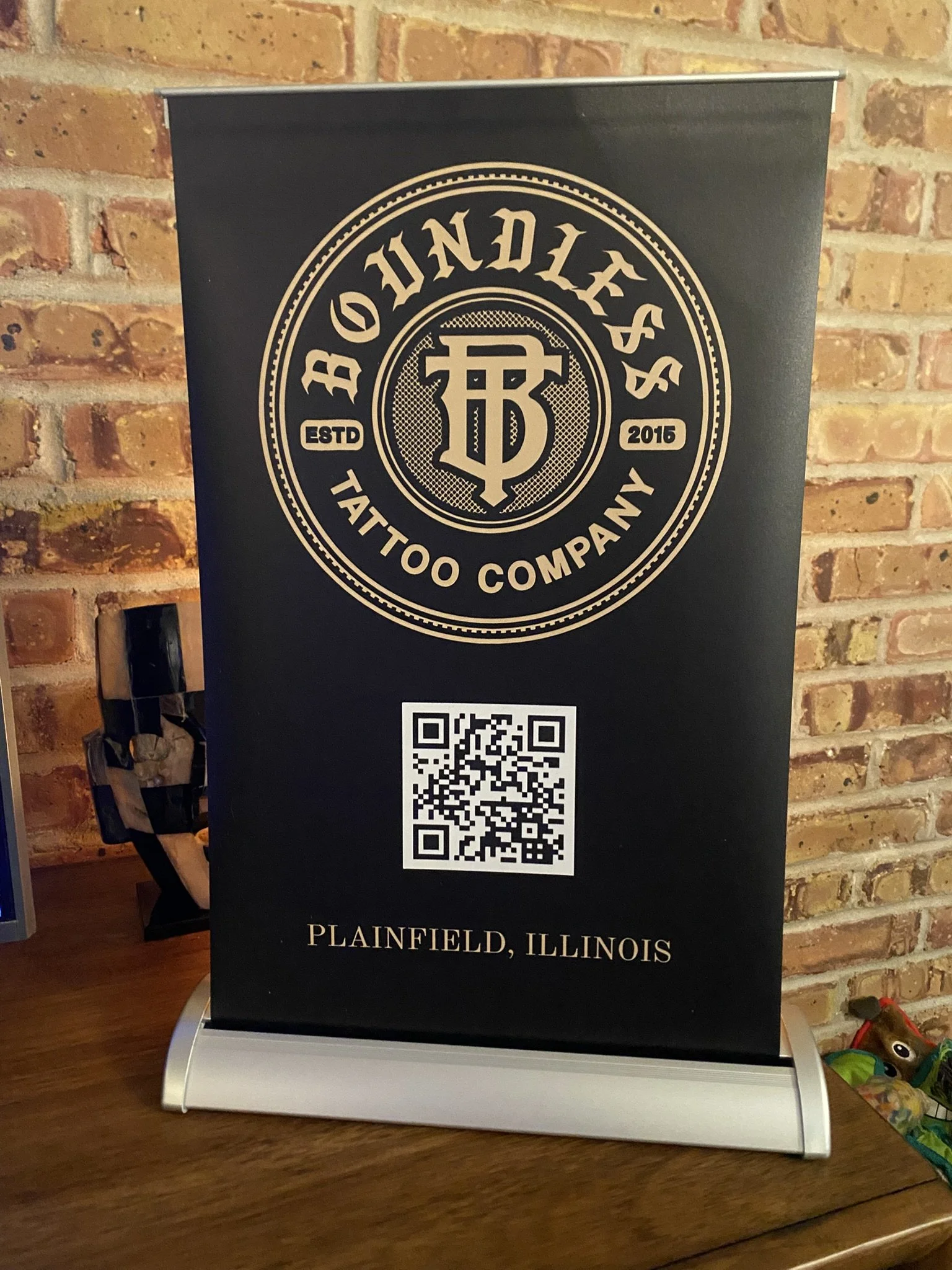 A promotional display for a tattoo company called 'Digdiff's Tattoo Company,' established in 2015, located in Plainfield, Illinois. The display features a black background with gold text and logo, a QR code, and is placed on a table against a brick wall.