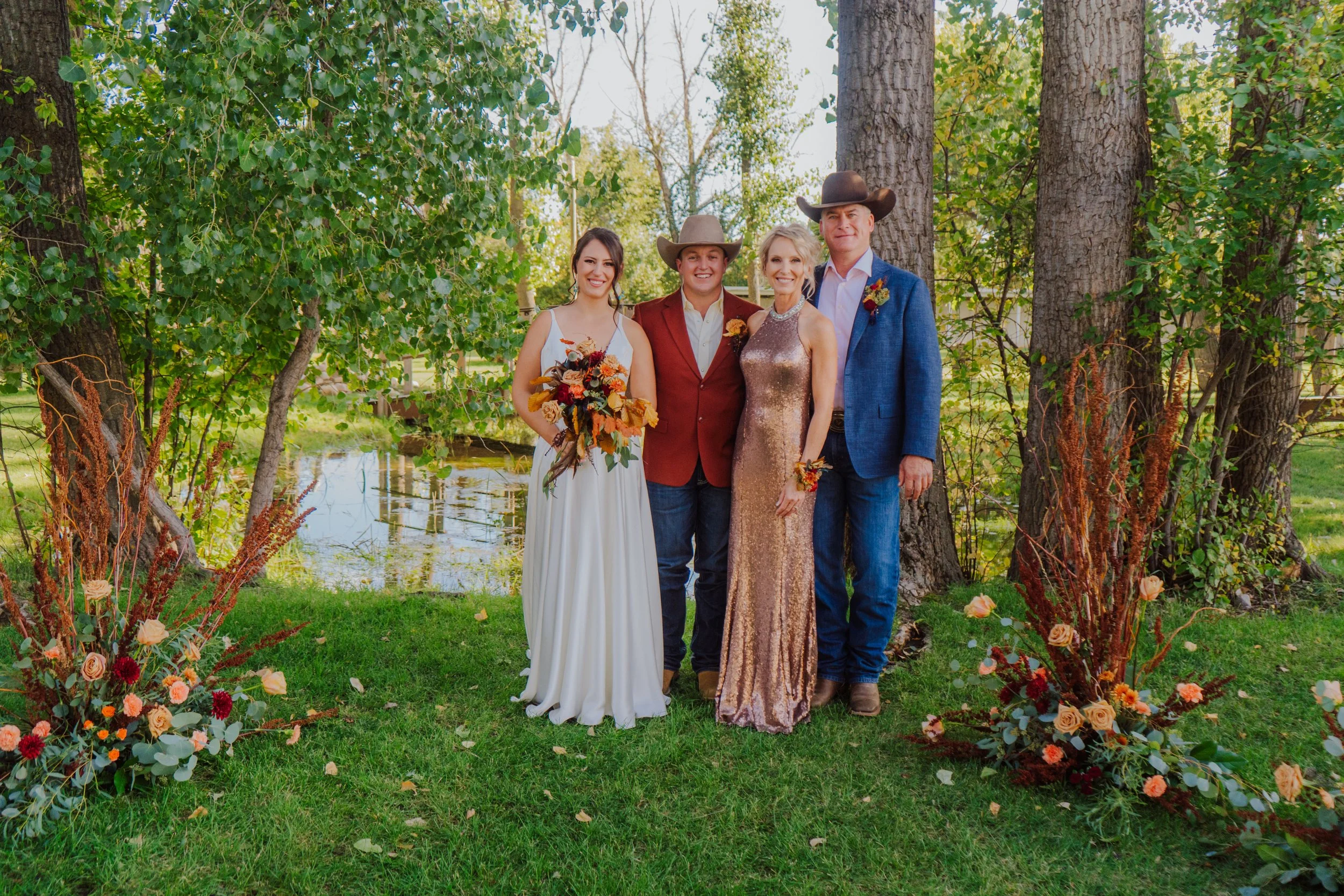 Group of four people dressed in formal attire standing outdoors near trees and a pond, posing for a photo at a wedding or special event.