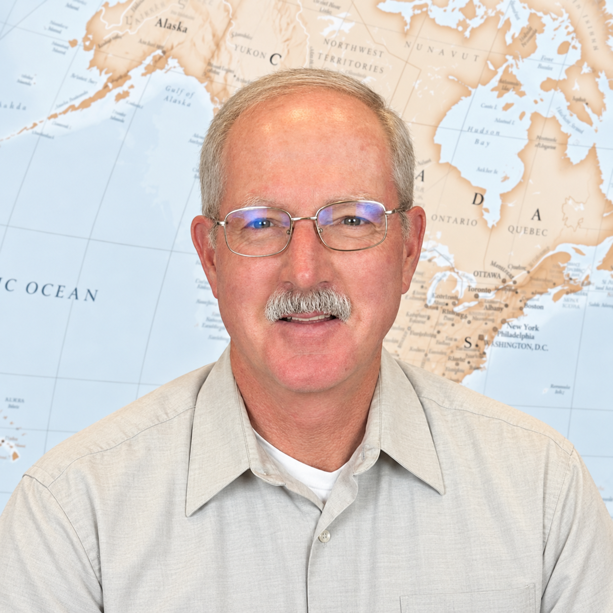 A middle-aged man with glasses and a mustache, wearing a light-colored button-up shirt, sitting in front of a bookshelf filled with books and binders.