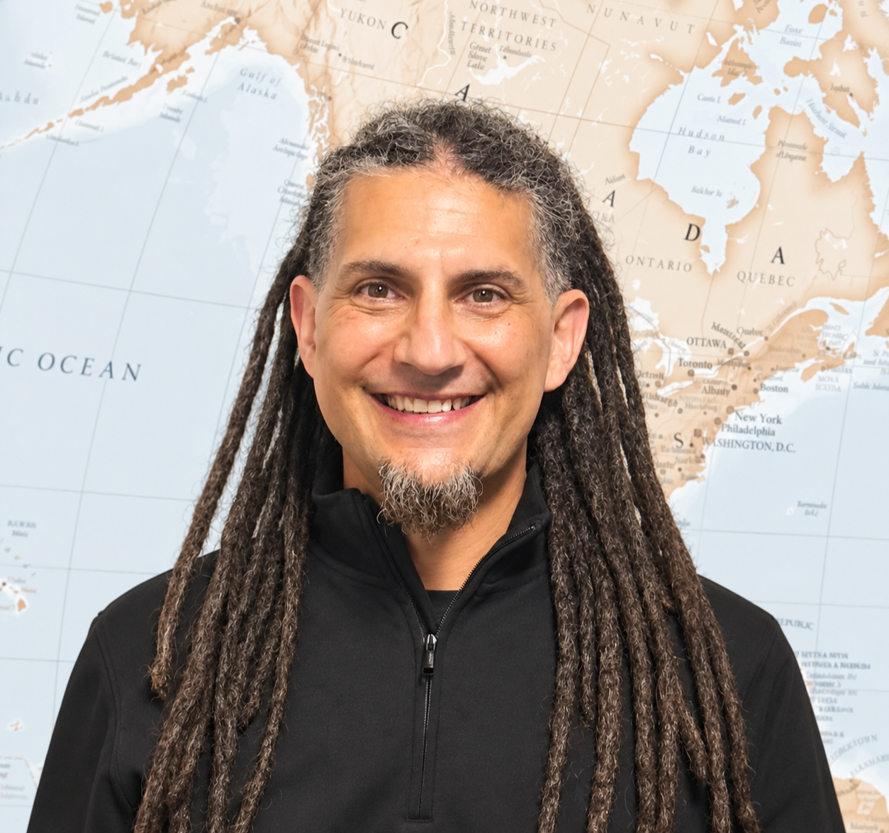 A smiling man with long dreadlocks, wearing a light green shirt, seated in front of a bookshelf filled with books.
