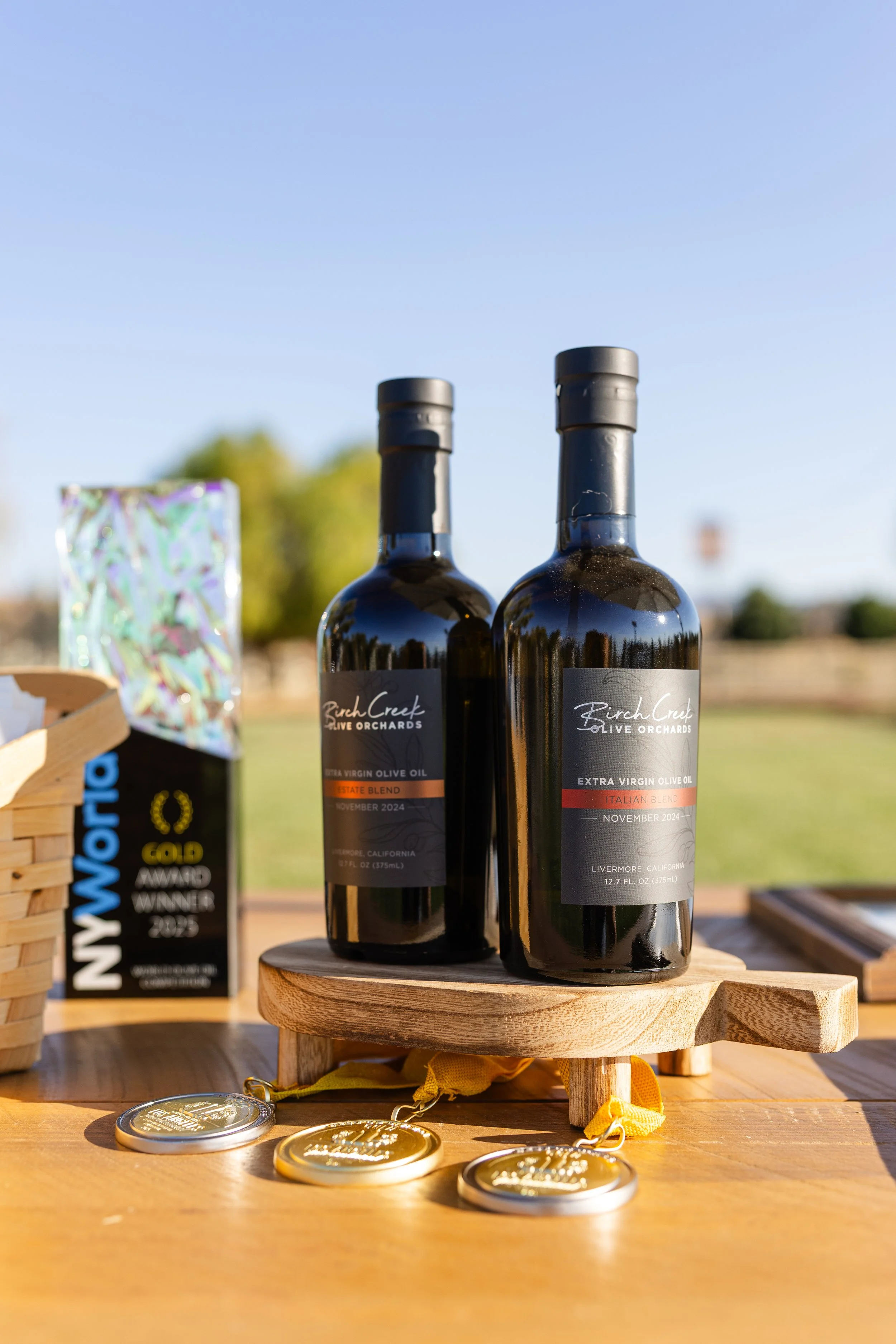 Two bottles of Birch Creek Olive Orchards extra virgin olive oil with labels indicating October and November 2024 in Livermore, California, placed on a small wooden stand on a table outdoors, with medals and a award plaque in the background.