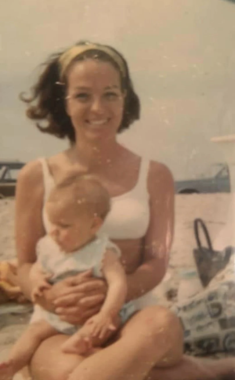 A smiling woman in a bikini cradles a baby in her arms, enjoying a sunny day by the water.