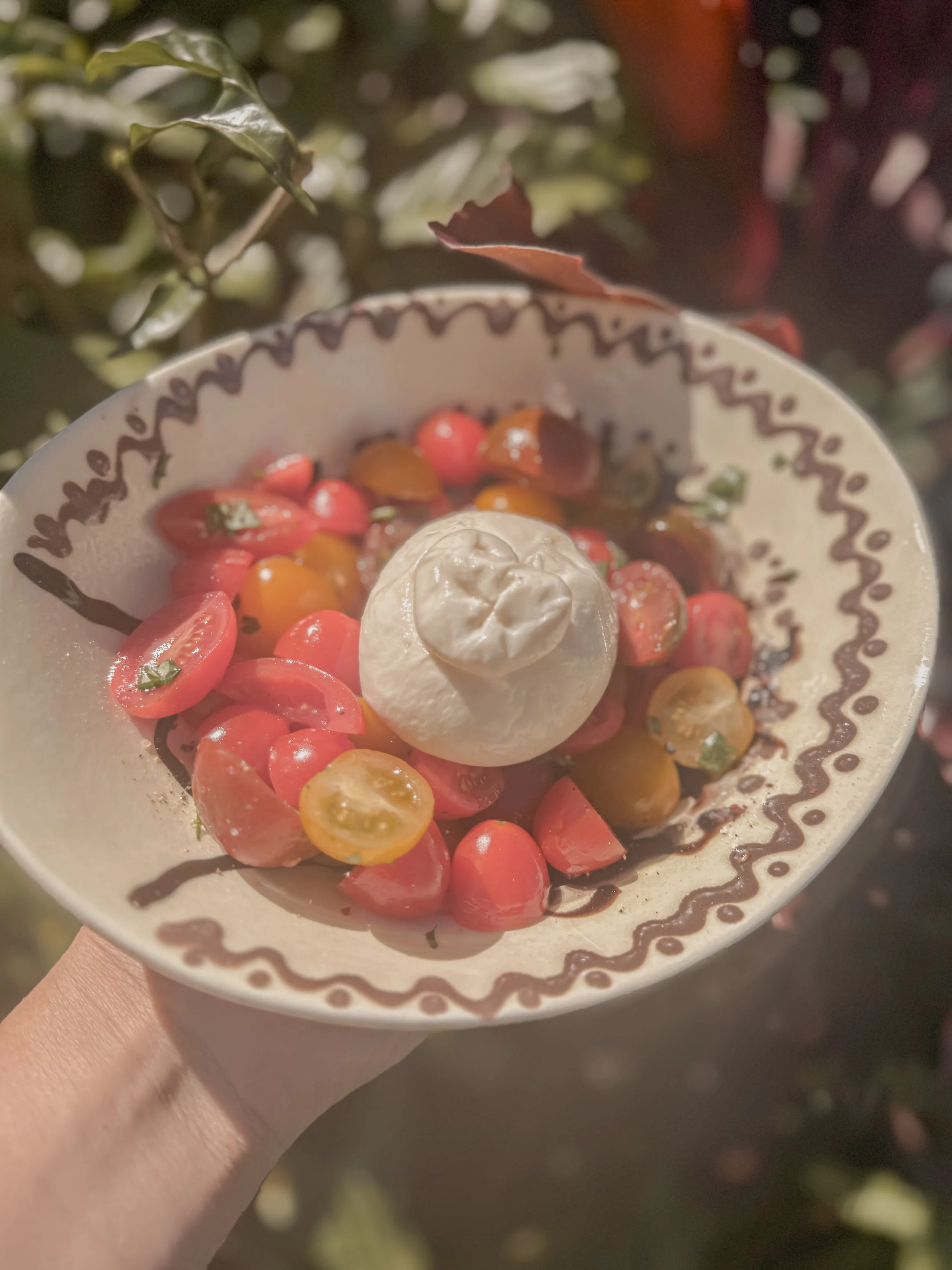 A bowl of cherry tomatoes with a dollop of creamy cheese or dressing on top, garnished with herbs, and decorated with a drizzle of balsamic glaze or similar dark sauce, held outside among green and reddish foliage.