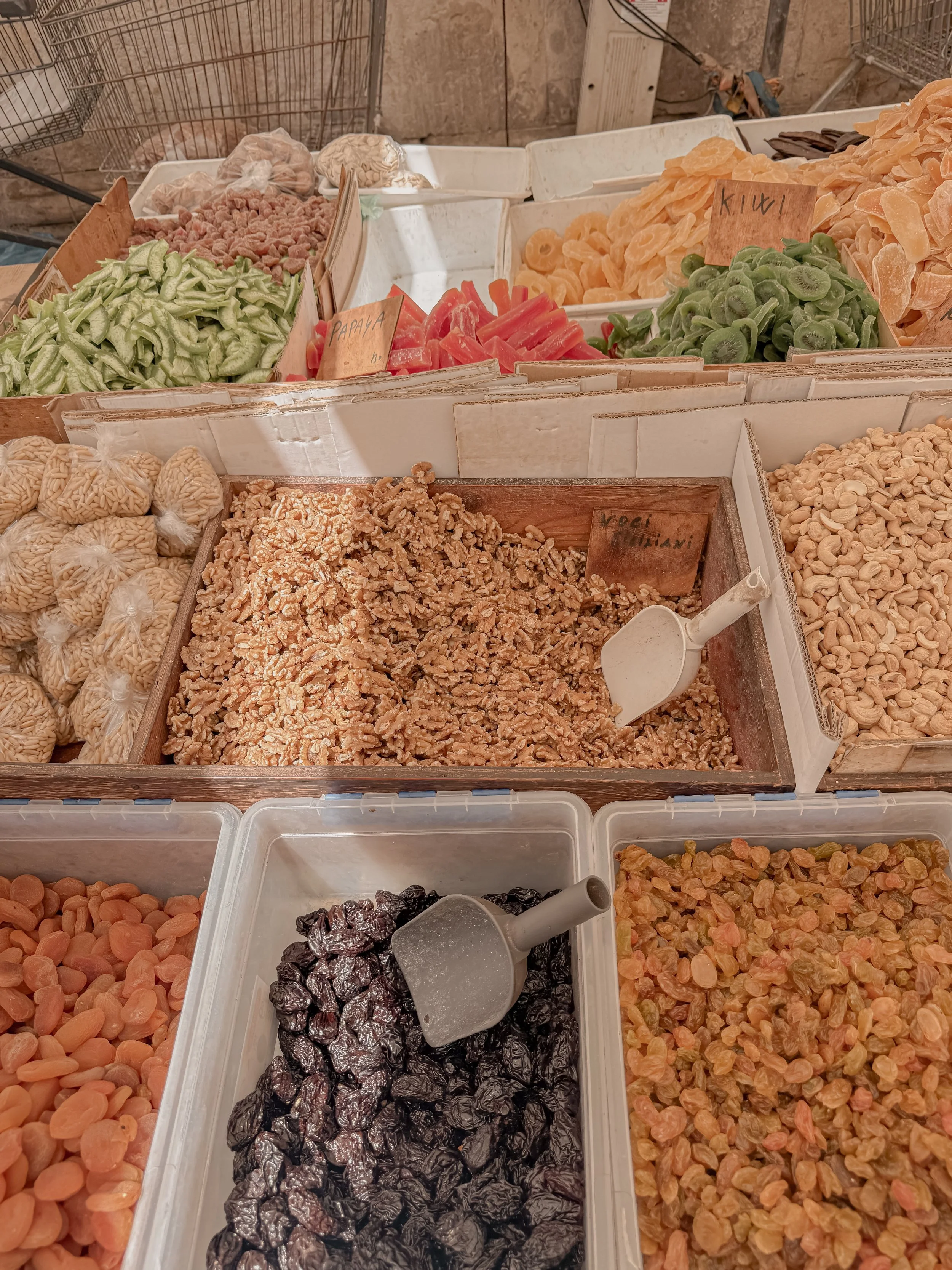 Variety of dried fruits and preserved foods displayed in white containers at a market, including dried apricots, dried prunes, dried figs, dried papaya, dried kiwi, and walnuts.