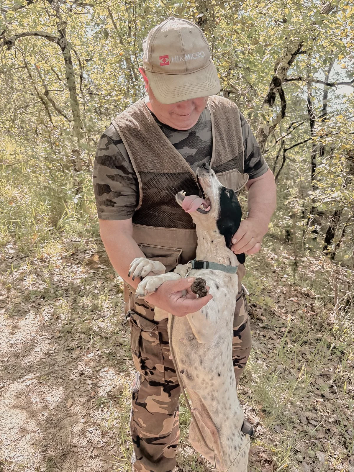 A man in camouflage clothing and a beige cap holding a large dog in a forested area. The dog is standing on its hind legs, looking up with its tongue out, and appears to be happy.
