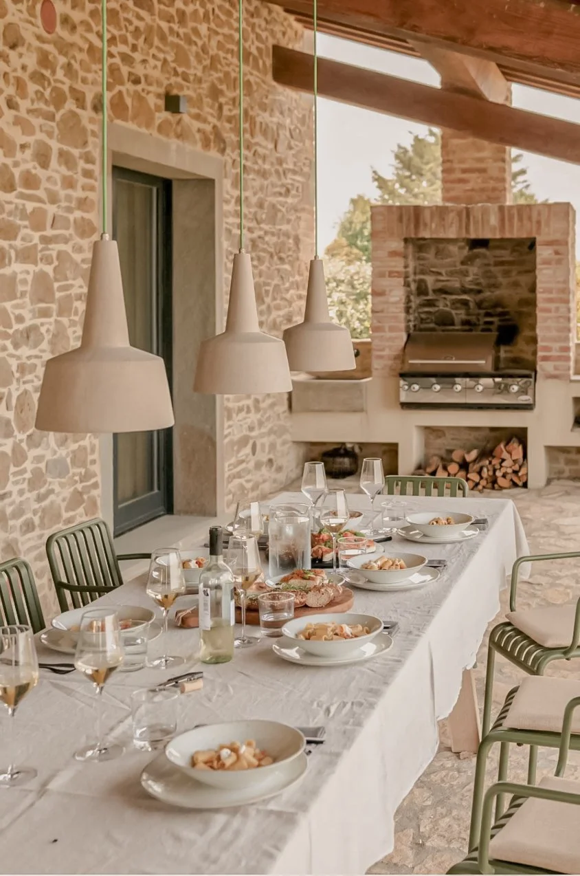 A dining table set for a meal with wine glasses, plates of food, and utensils, in a rustic outdoor space with stone walls, a brick fireplace, and hanging pendant lights.