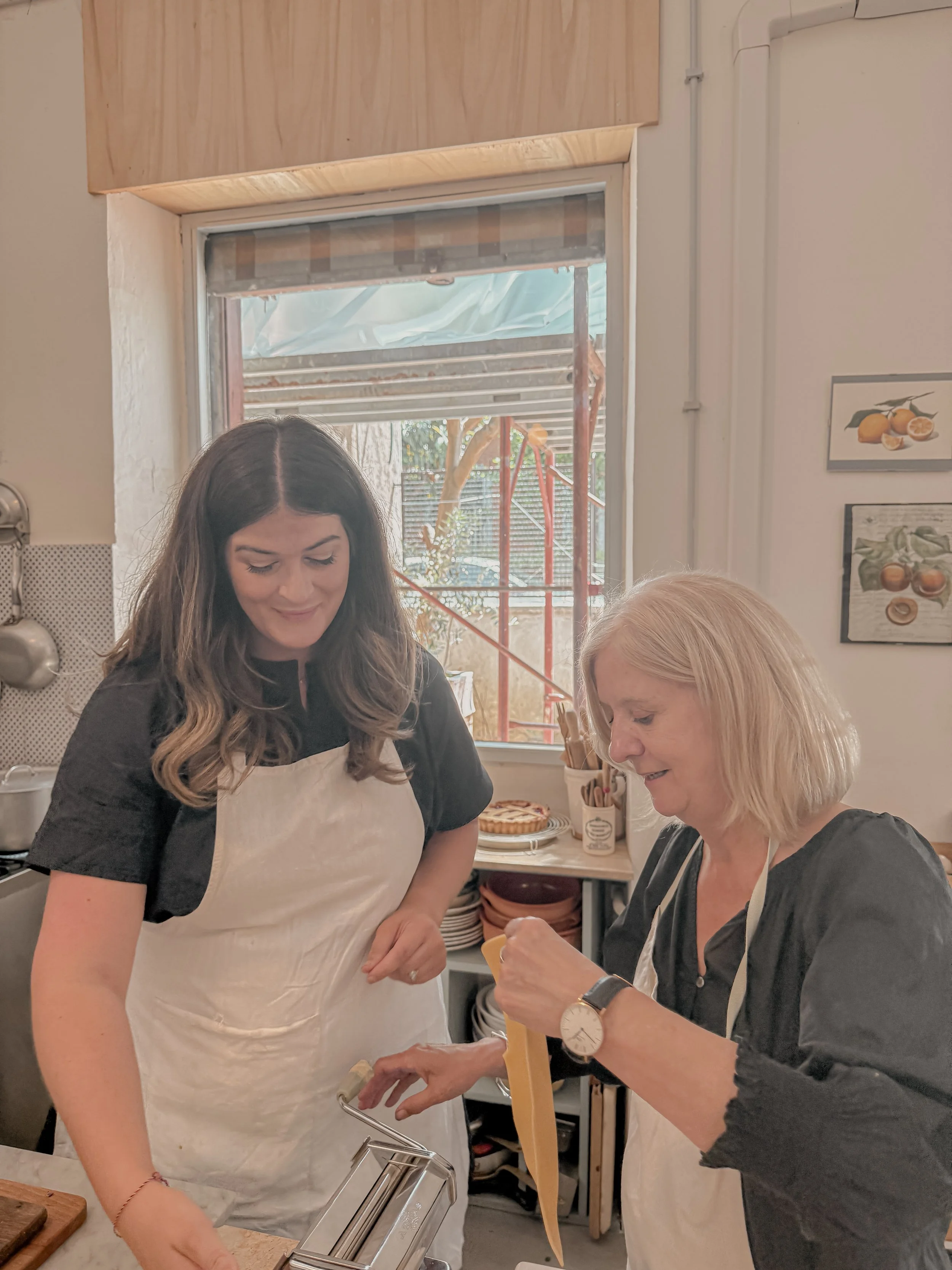 Two women making pasta in a kitchen, one younger with dark hair, one older with blonde hair, both wearing aprons, with a window showing an outdoor scene.