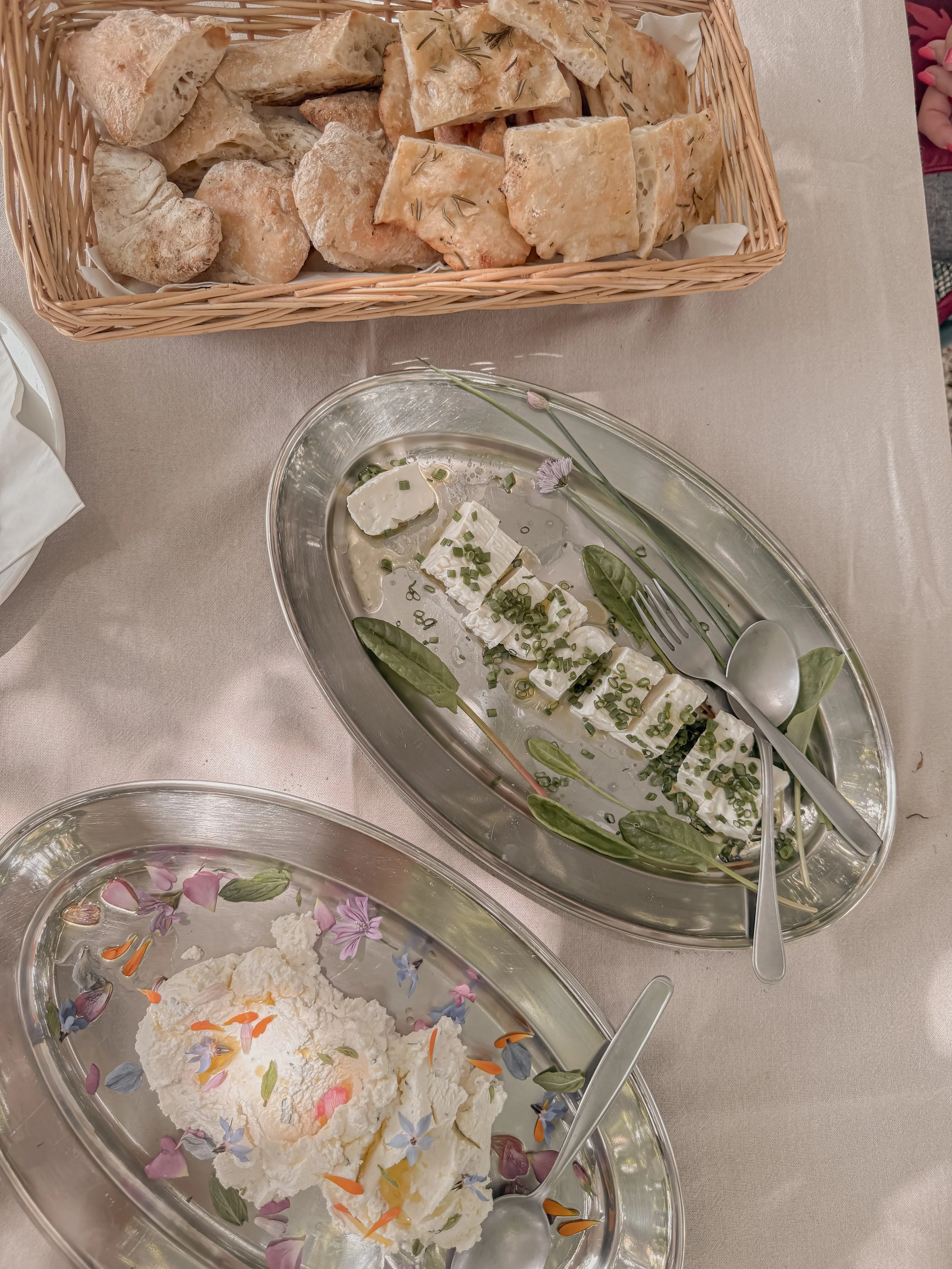 A basket of sliced focaccia bread, a bowl of sliced cheese garnished with chives and herbs with edible flowers, and a bowl of ricotta cheese with edible flower petals on a table.