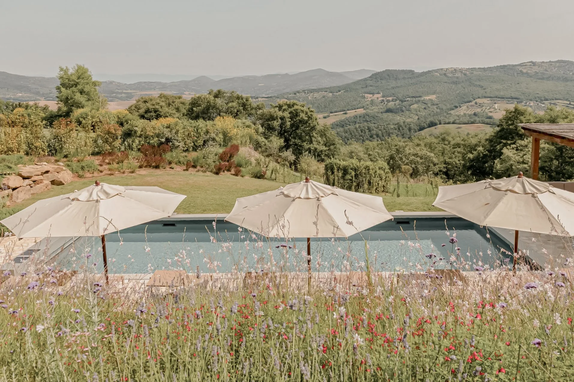 Swimming pool with three white umbrellas in a grassy yard, overlooking rolling green hills and mountains under a cloudy sky.