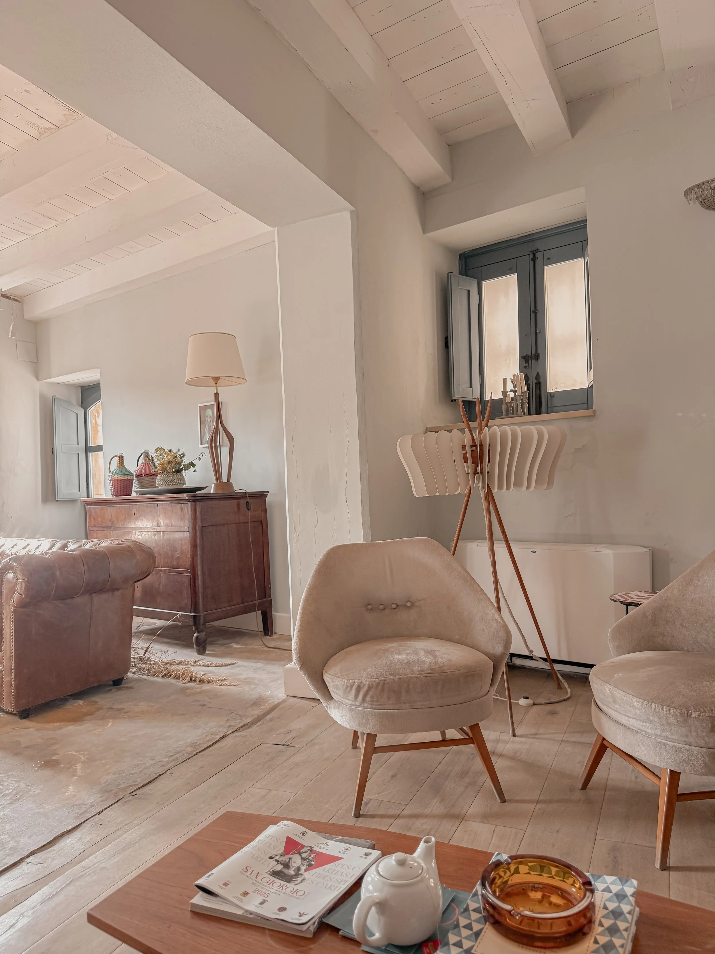 Living room with beige chairs, a wooden coffee table with magazines and a teapot, a vintage wooden cabinet, a tall floor lamp, and a window with gray shutters.