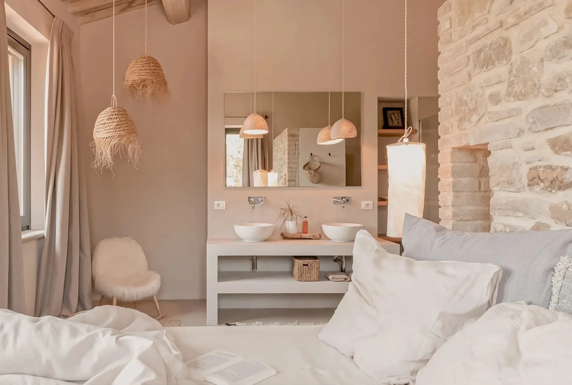 A cozy bedroom with a double bed, white bedding, pillows, and an open book. Behind the bed is a rustic stone wall. To the left, there are curtains, a window, and a fluffy chair. In the background, a vanity area with two white vessel sinks, a mirror, pendant lights, and shelves with decor is visible.