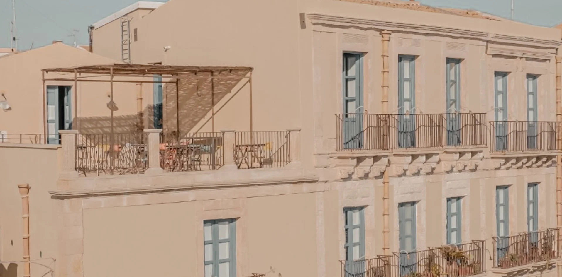 Buildings with multiple windows and balconies in a warm-colored cityscape, including a terrace with outdoor furniture and a lattice-style roof structure.