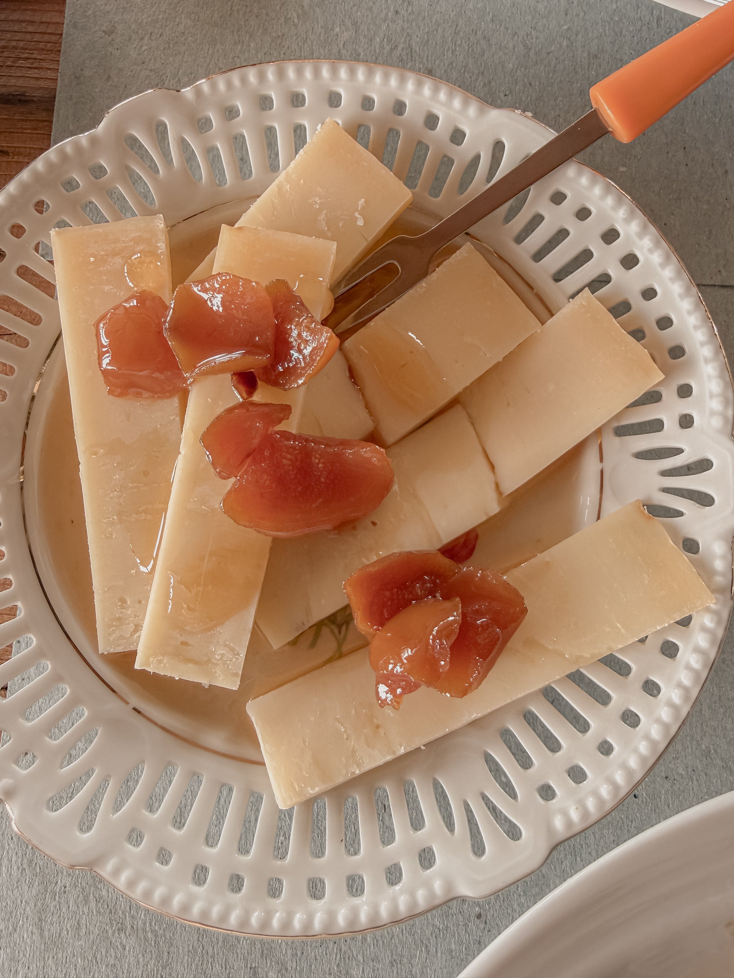 A white decorative plate with slices of hard cheese and pieces of dried apricots on a gray table.
