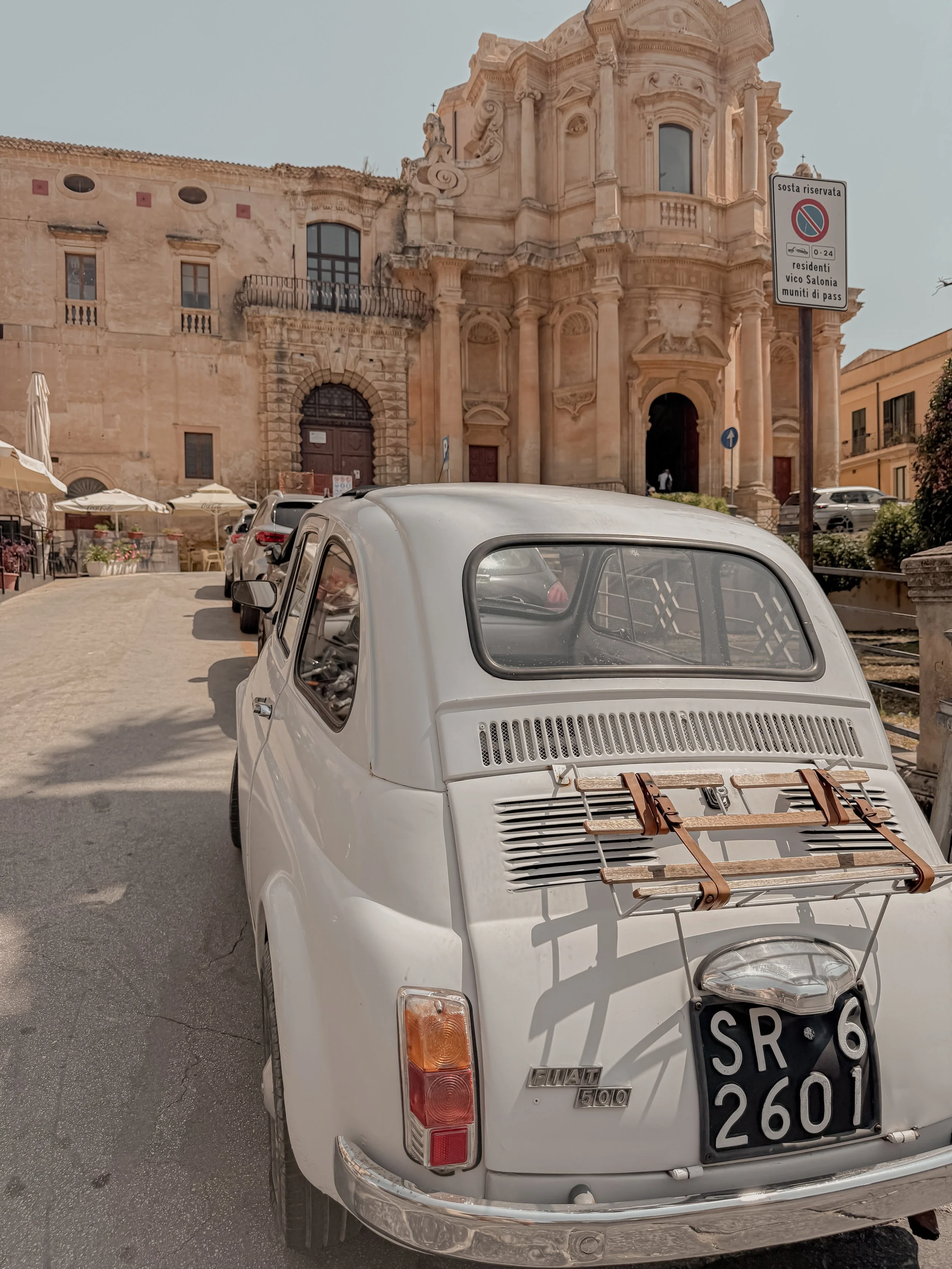 A vintage white Fiat 500 car is parked on a street in front of a historic Baroque church with ornate architecture. The car has a luggage rack on the back and a black license plate with white characters.”}