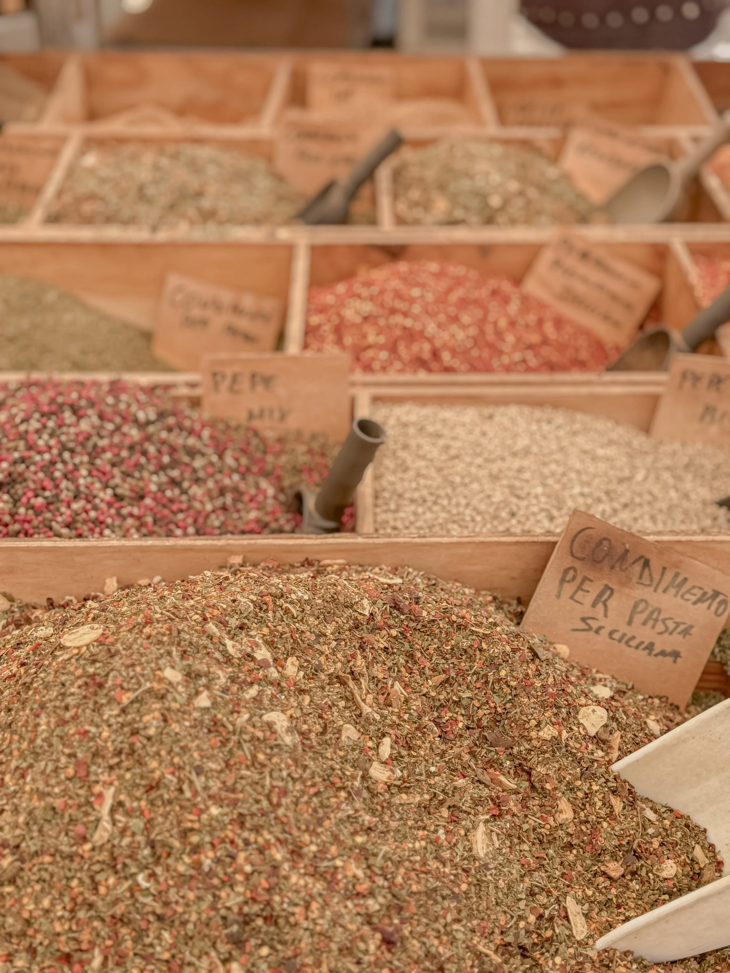 A display of various dried spices and herbs in wooden bins at a market, with small handwritten signs indicating the spice names.