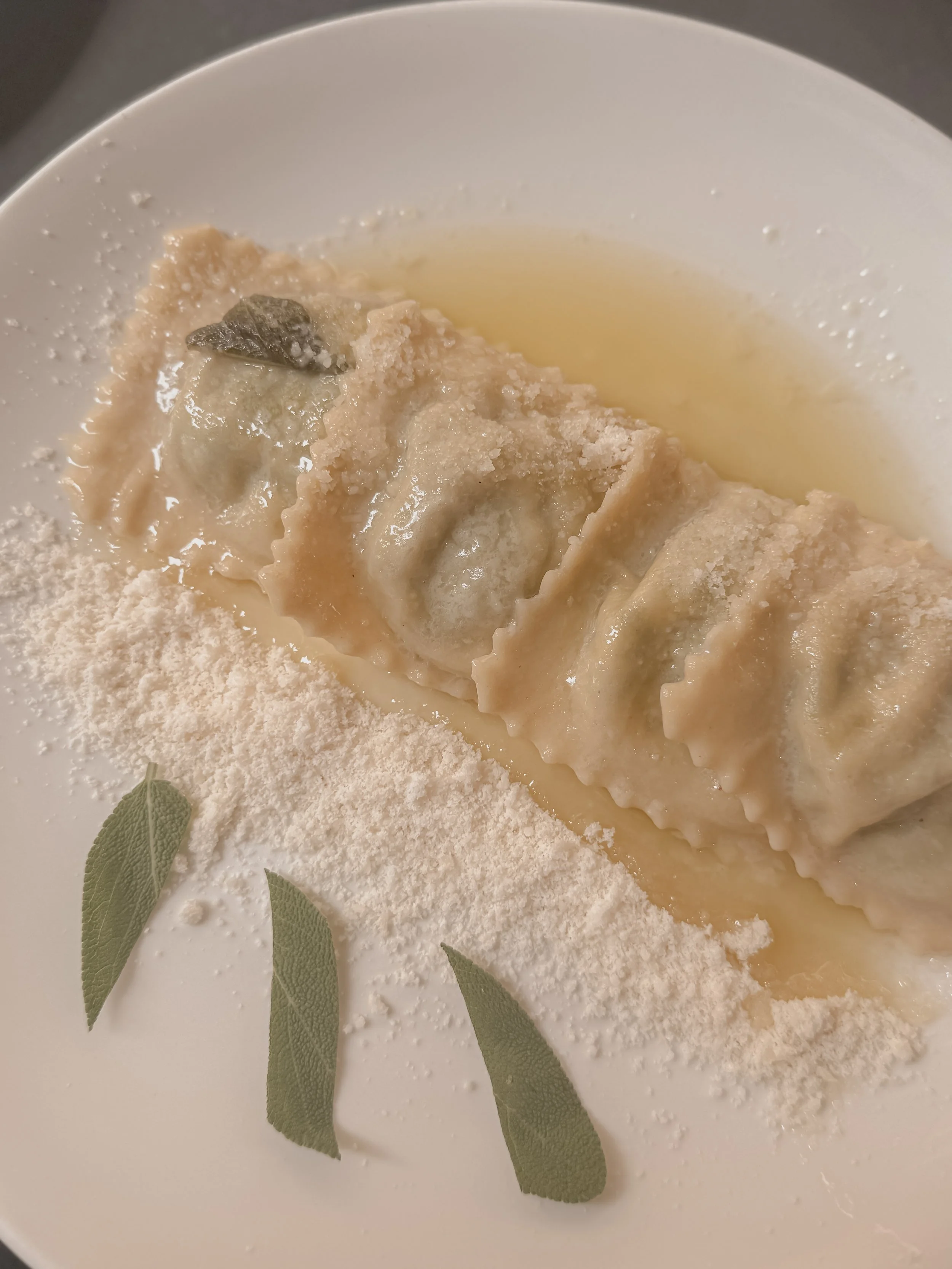A white plate with cooked ravioli pasta topped with grated cheese and sage leaves, served with a light sauce.