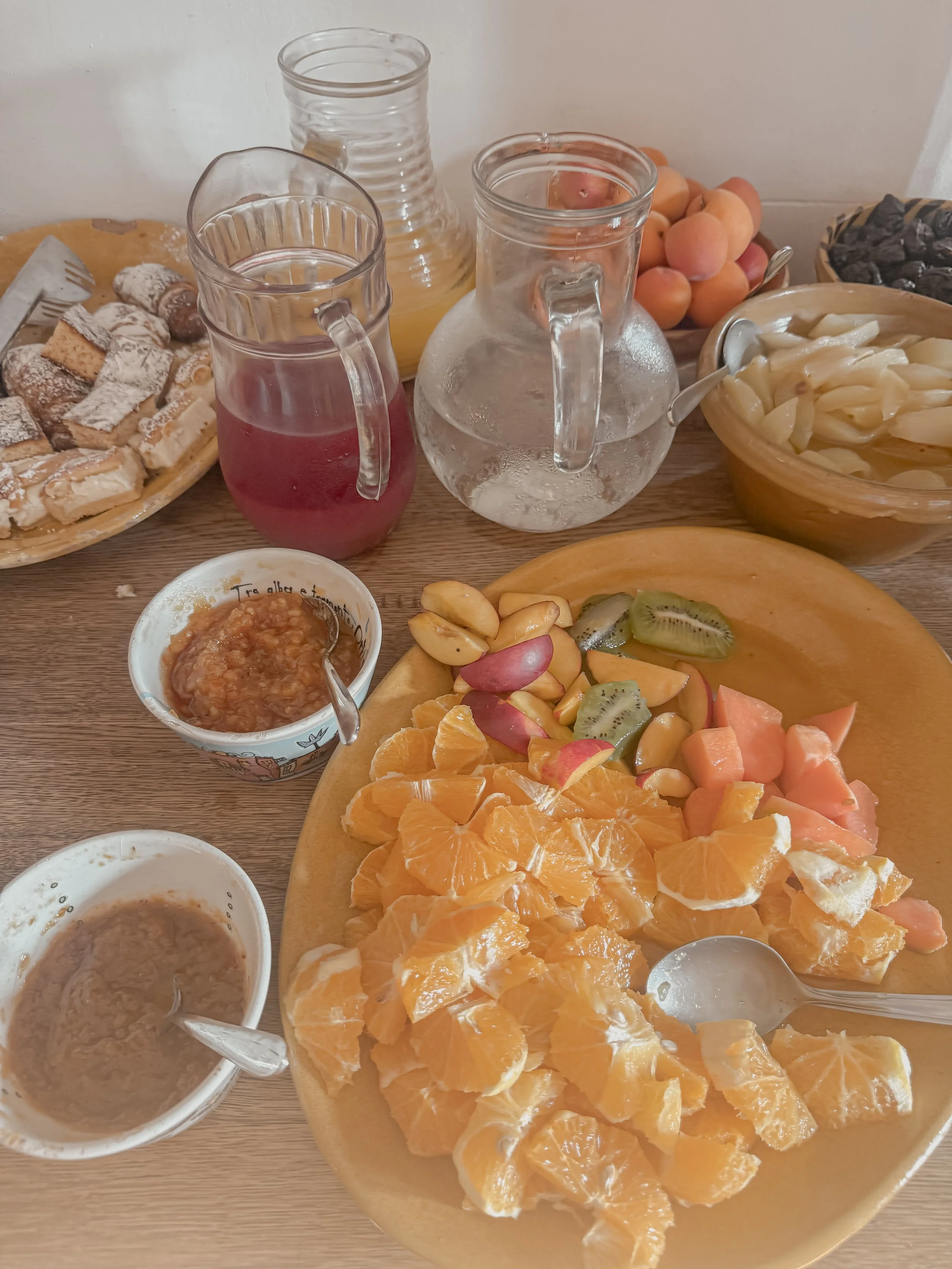 Table set with various breakfast foods including pitchers of juice, a bowl of peaches and black olives, a plate of pastries, and a platter of fresh fruit such as orange slices, kiwi, apple slices, and melon.
