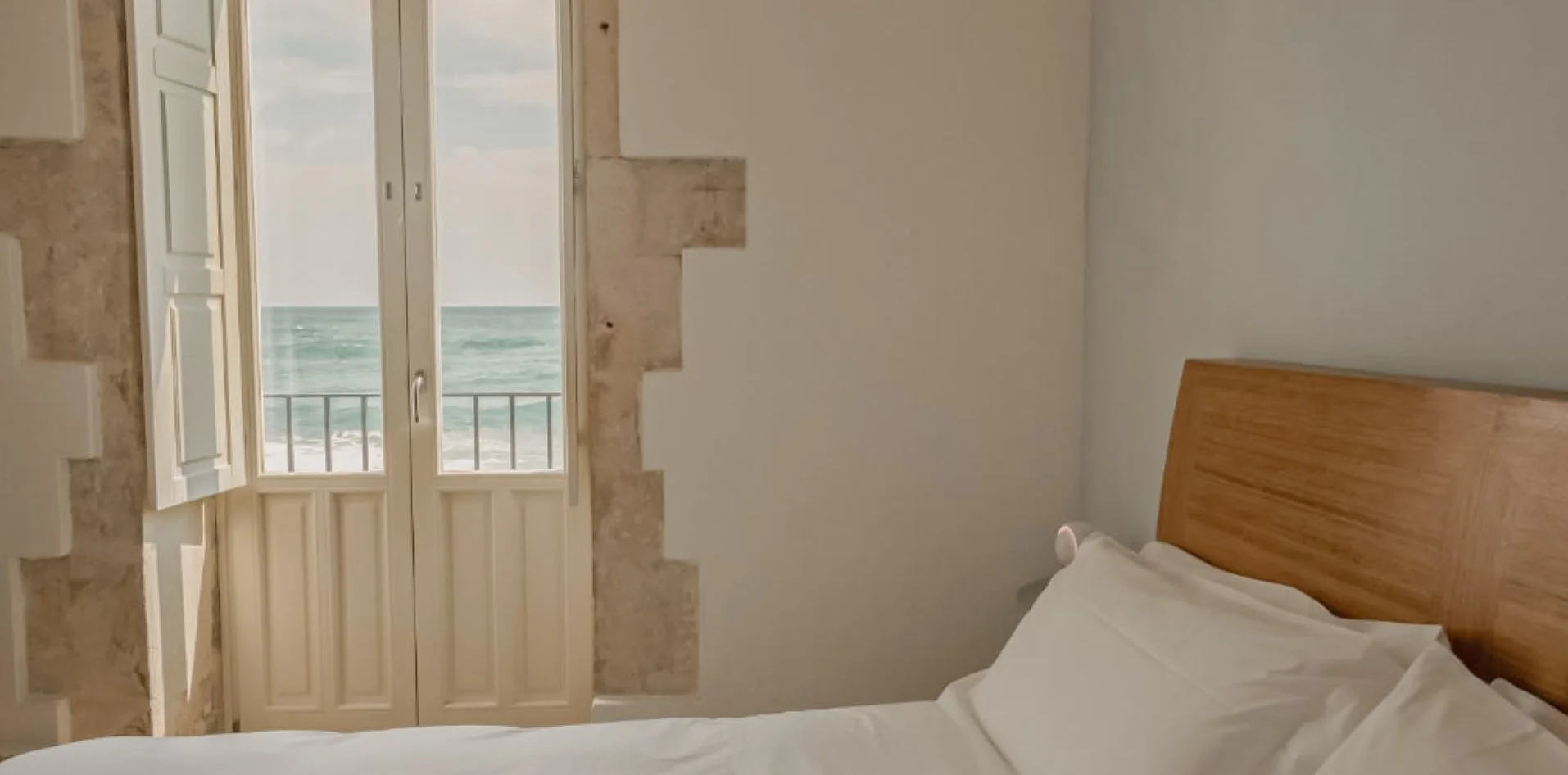 View of a bedroom with a bed, a wooden headboard, and an open French door leading to a balcony with an ocean view
