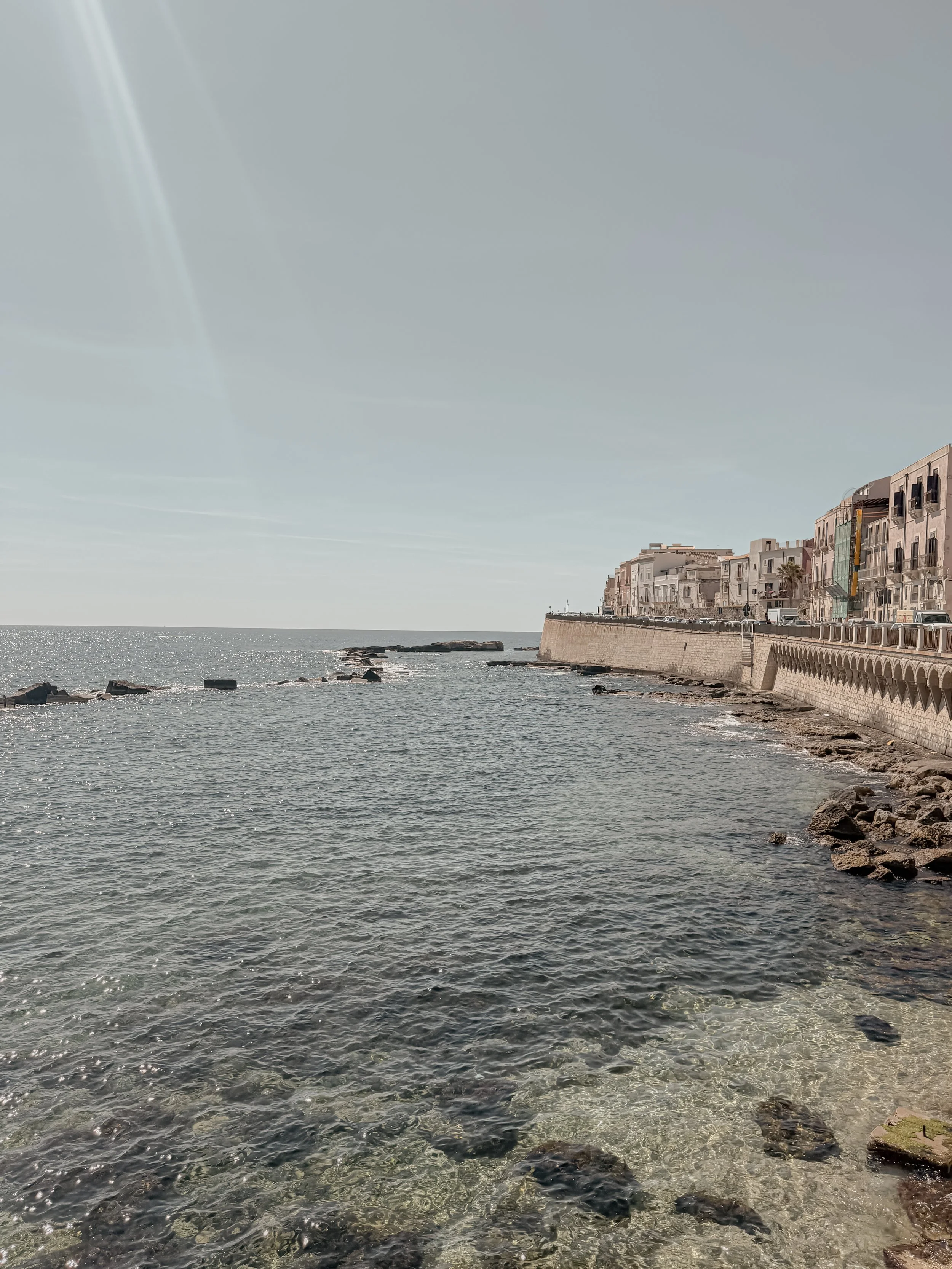 A coastal scene with a row of buildings along the shoreline, a concrete promenade, and a calm sea with rocks in the water, under a clear sky.