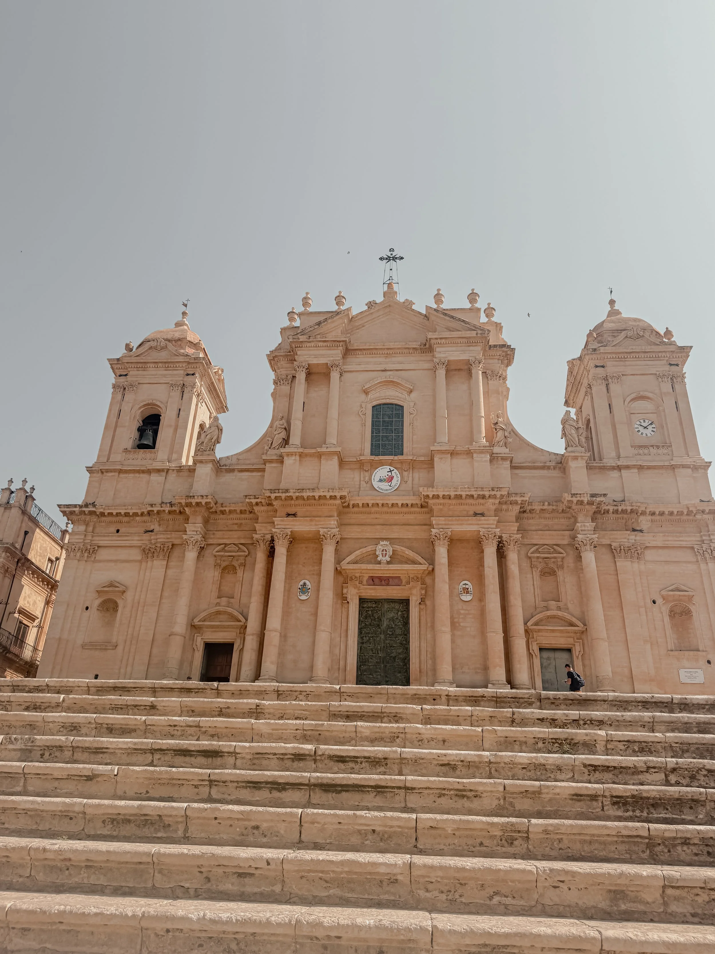 A large, historic church with a classical architectural style, featuring two towers, stone steps leading up to the entrance, and a small person sitting on the steps.