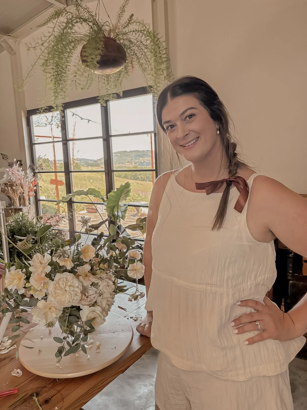 A woman smiling and taking a selfie in a bright room with large windows, floral arrangements, and plants, during daytime.