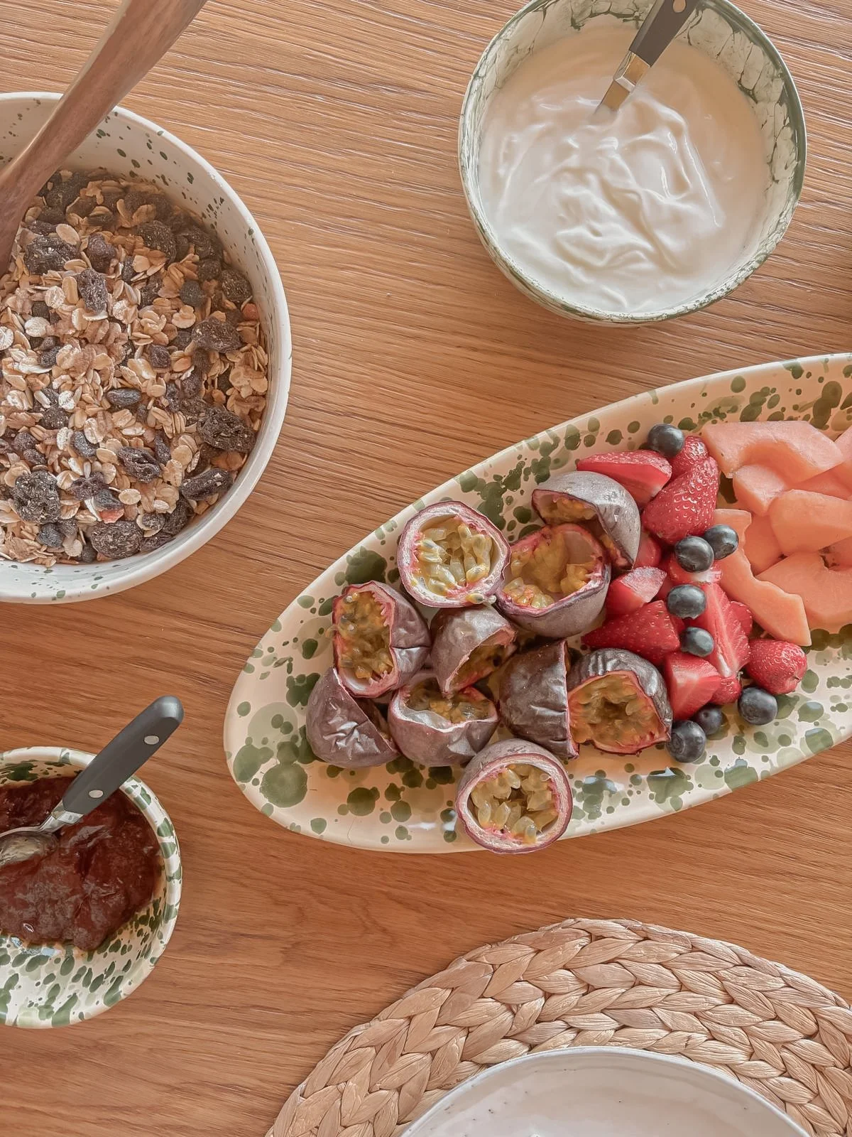A breakfast table with a bowl of granola, a bowl of yogurt, a plate of fresh mixed berries, and a small bowl of jam on a wooden table.