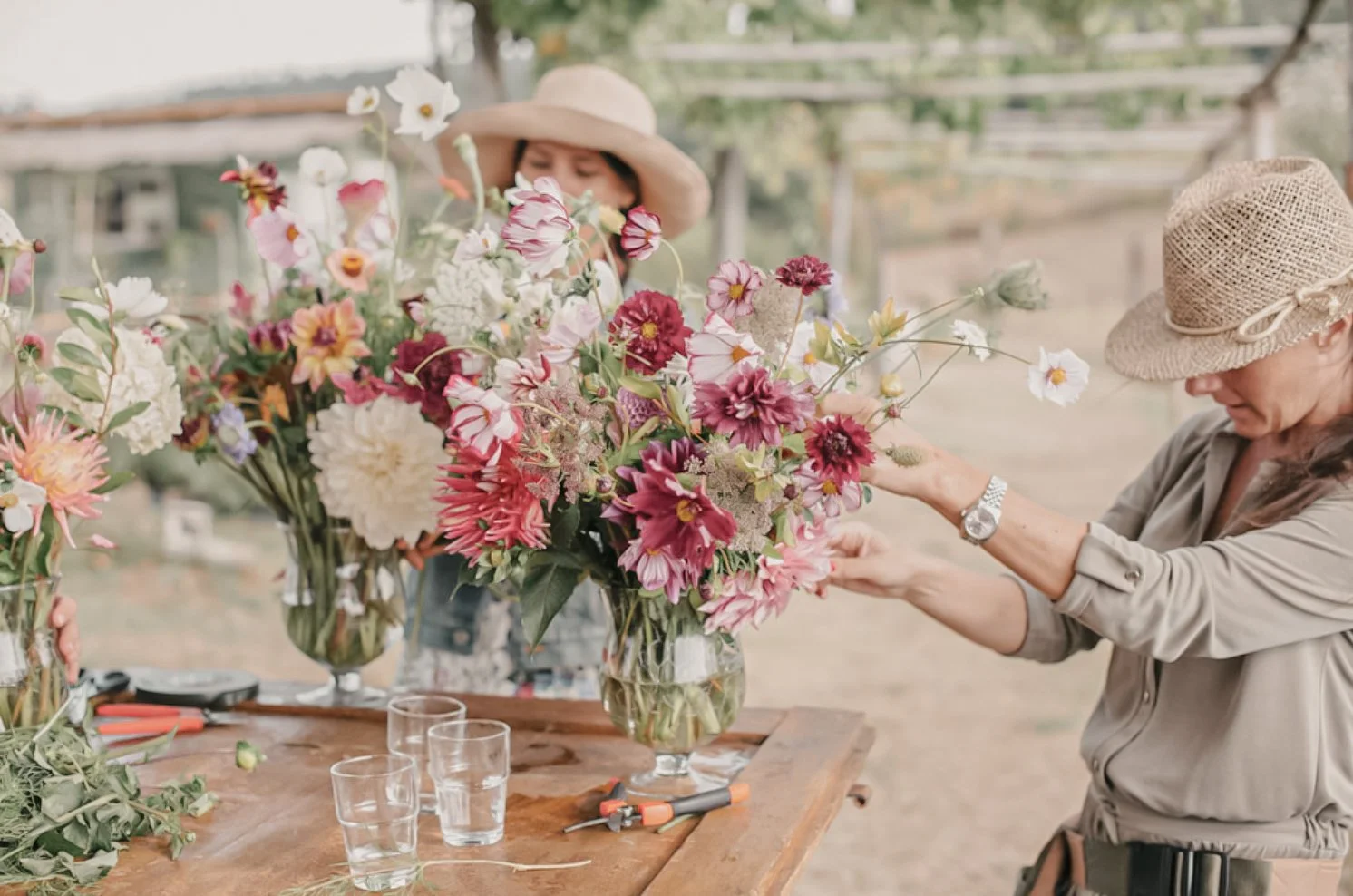 Two women arranging colorful flowers in vases on a wooden table outdoors, wearing sun hats and casual clothing.