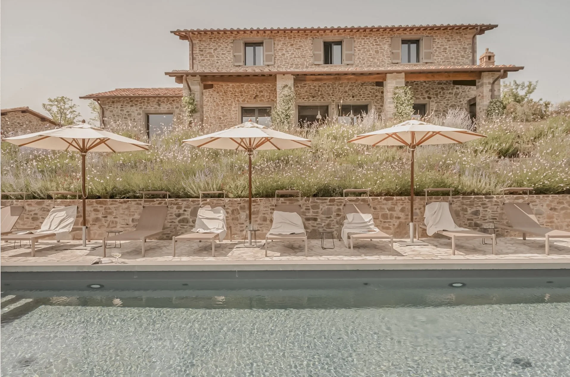 Poolside view of a stone house with large windows, surrounded by lounge chairs and umbrellas, view from pool level.
