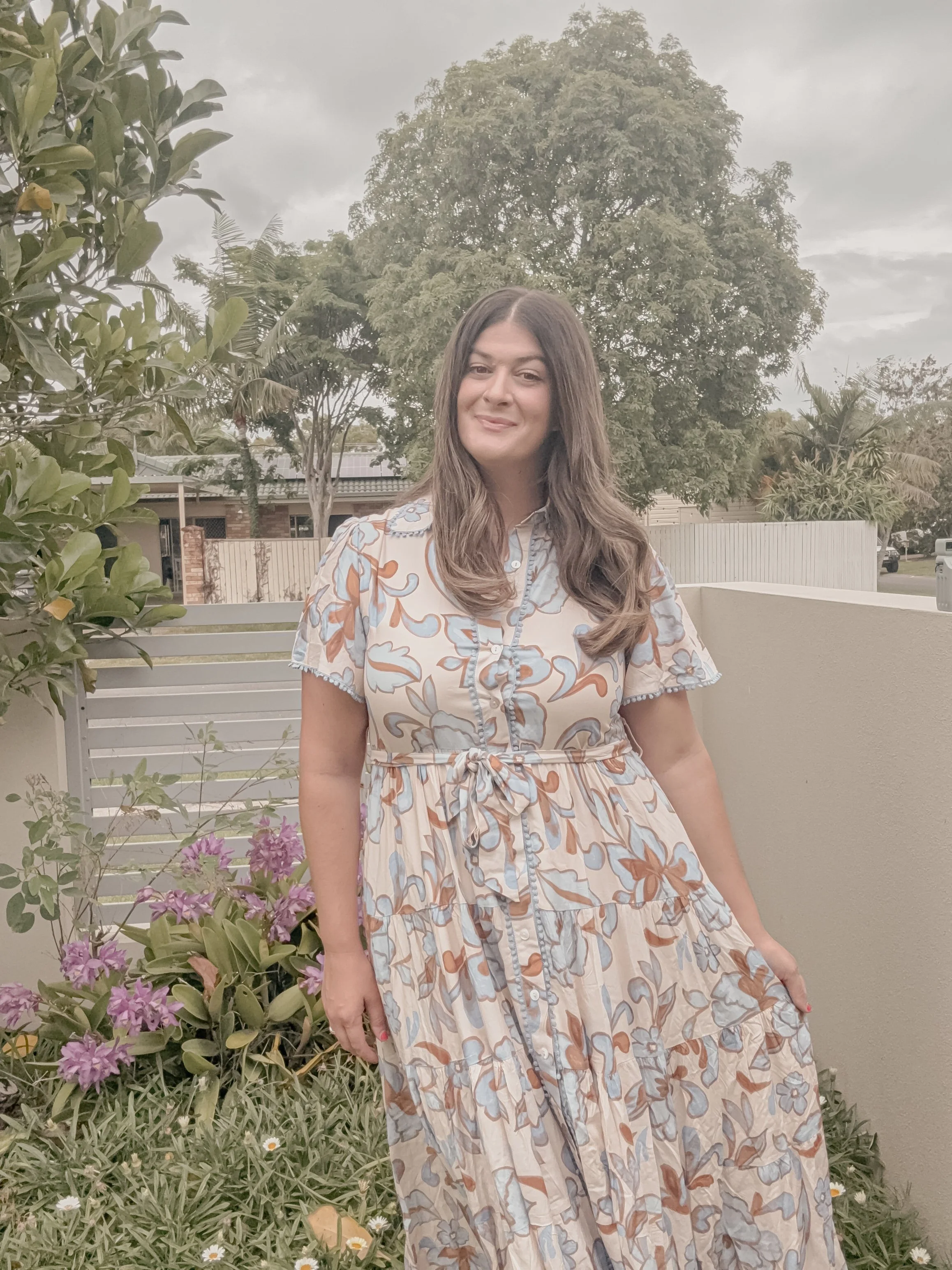 A woman with long brown hair smiling in a garden, wearing a flowy, floral patterned dress with short sleeves and a tied waist, standing beside a bush with purple flowers and green leaves.