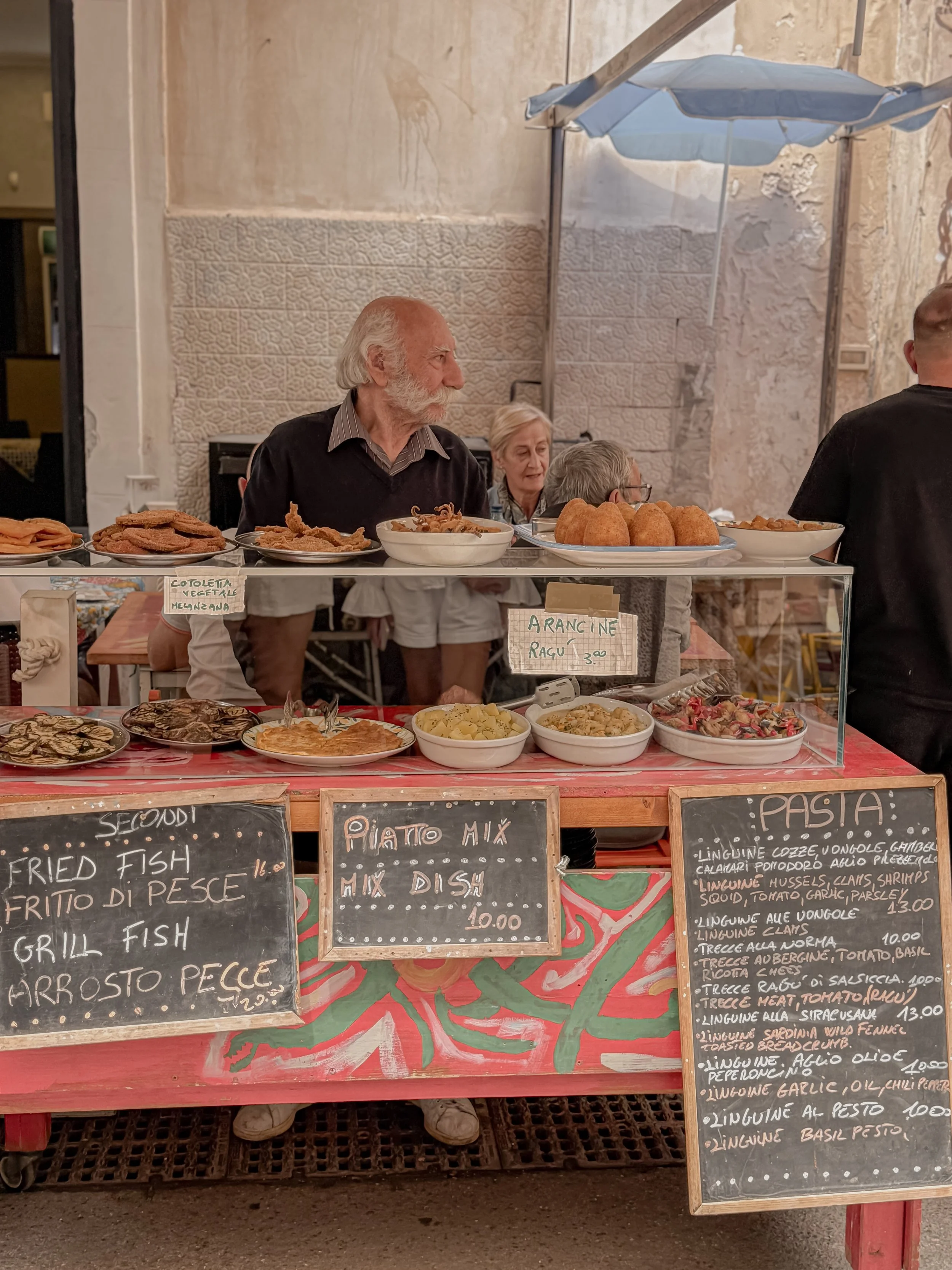 Street food stall with various Italian dishes on display, including fried fish, pasta, and sauces, with handwritten menu signs in Italian and English.