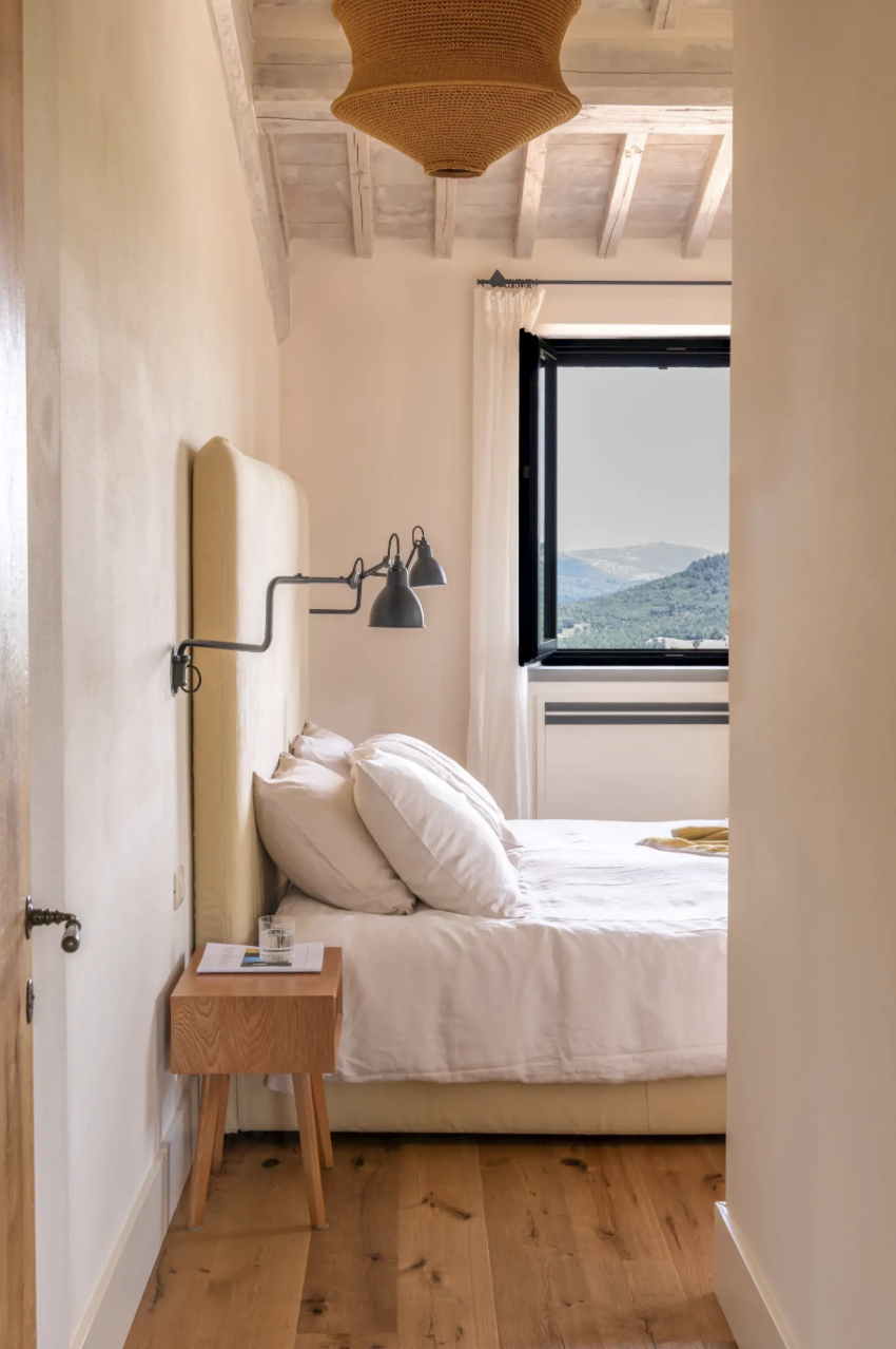 A cozy bedroom with a bed, cream-colored headboard, and white bedding. There is a small wooden bedside table with a glass of water and magazines. A window shows scenic mountains outside, and a beige ceiling lamp hangs from the wooden ceiling.