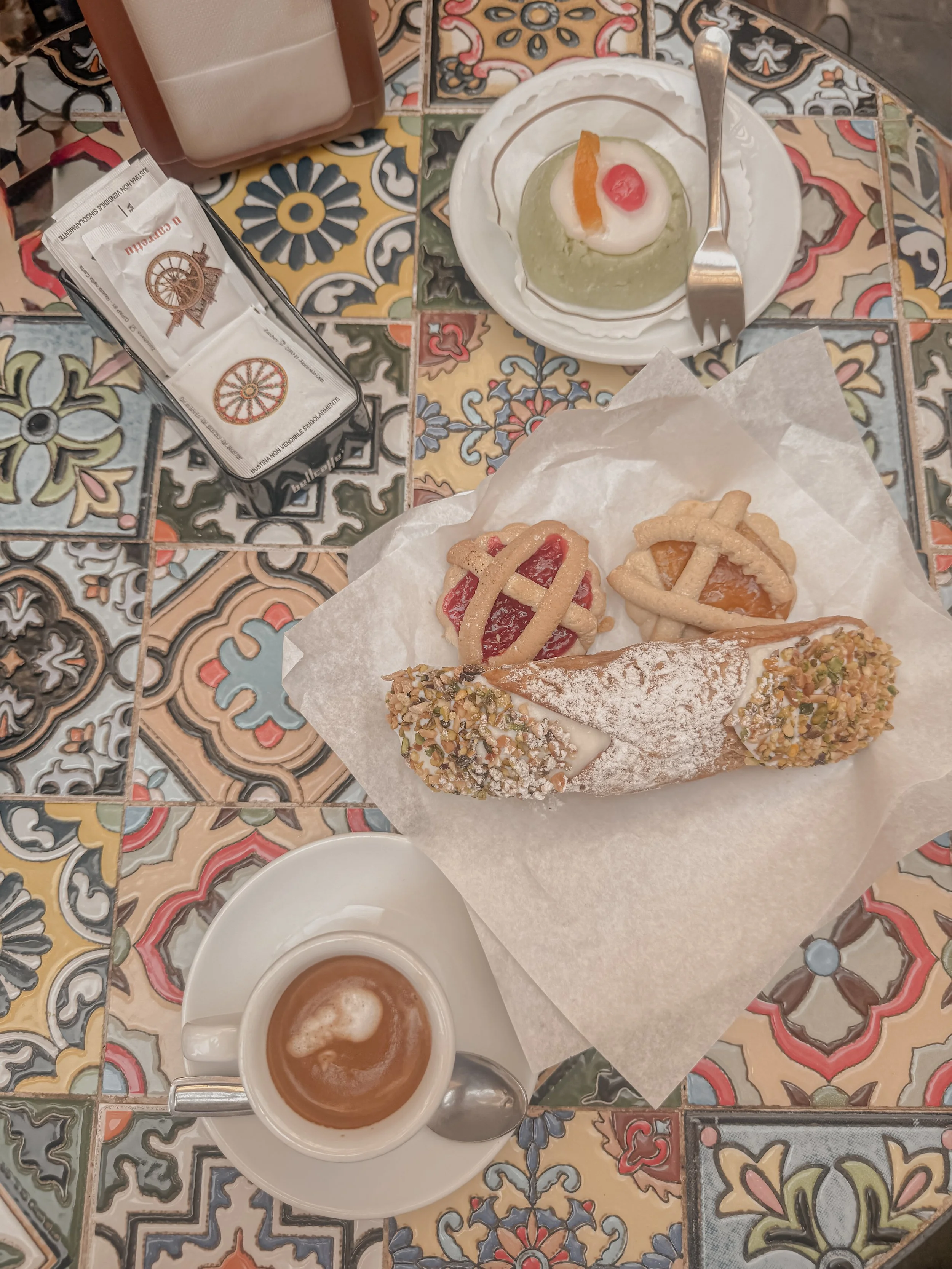 A colorful tiled table with a cup of coffee, a slice of cannoli, and a small plate with fruit and pastry, along with a package of sugar and a container of milk.