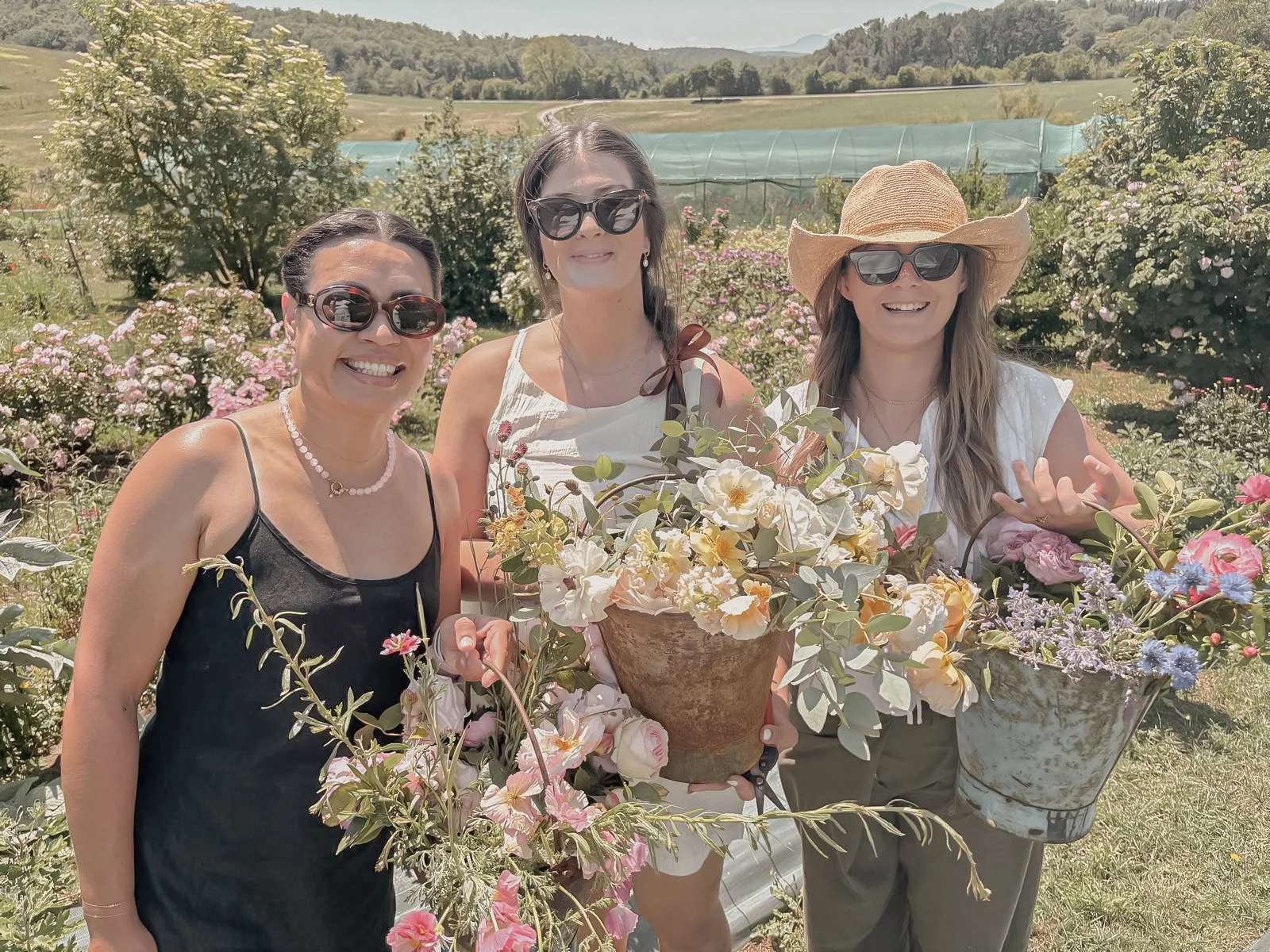 Three women smiling and holding flower baskets in a garden with blooming flowers and greenery, wearing sunglasses and summer clothes.