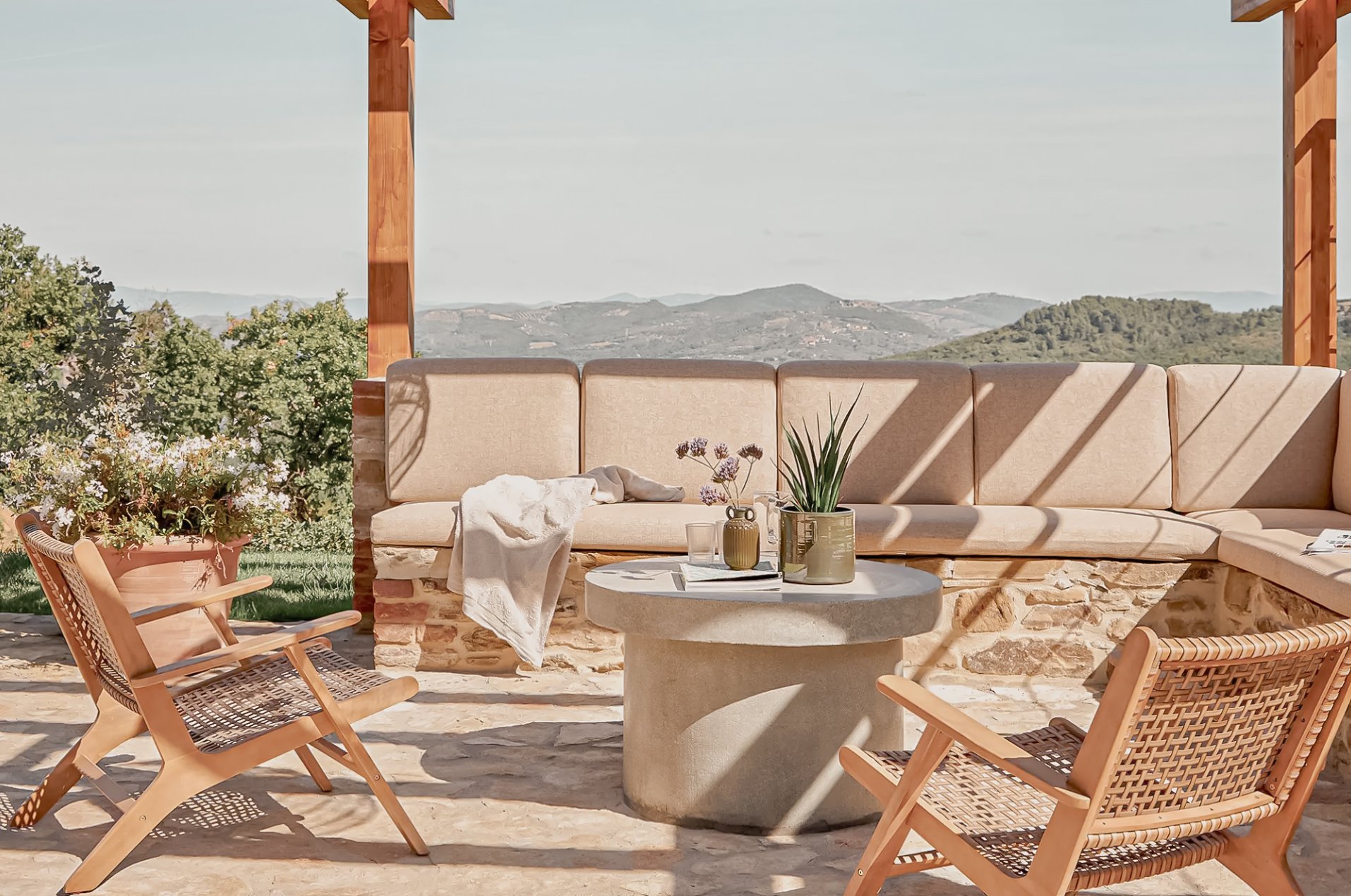 Outdoor seating area with a stone and wooden structure, beige cushioned bench, two wooden chairs with woven seats, a round concrete table with potted plants and glasses, and scenic view of hills and trees in the background on a sunny day.