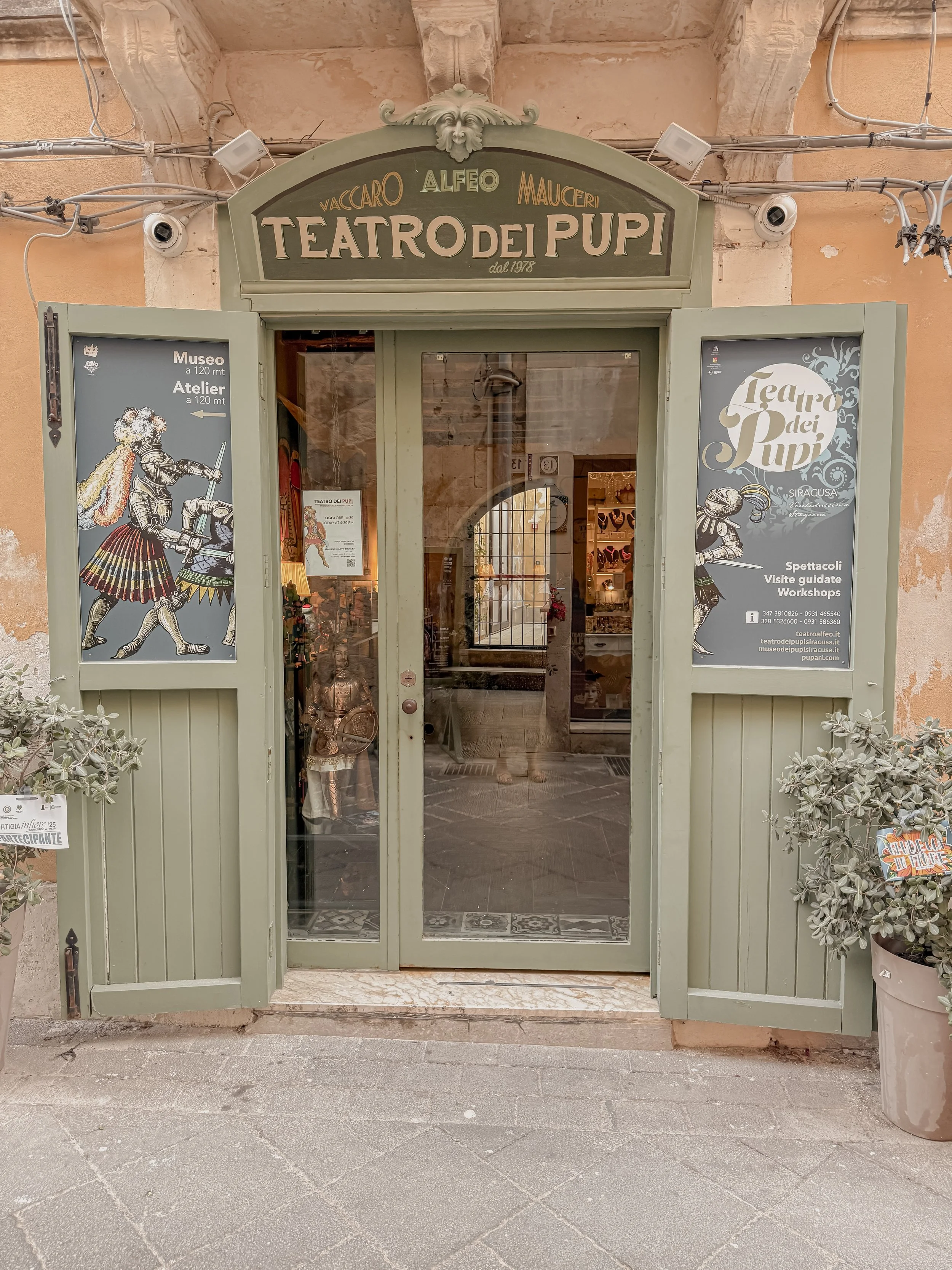 Entrance to Teatro dei Pupi, a puppet theater in Siracusa, Italy, with green wooden doors, decorative sign, and posters advertising shows and guided tours.