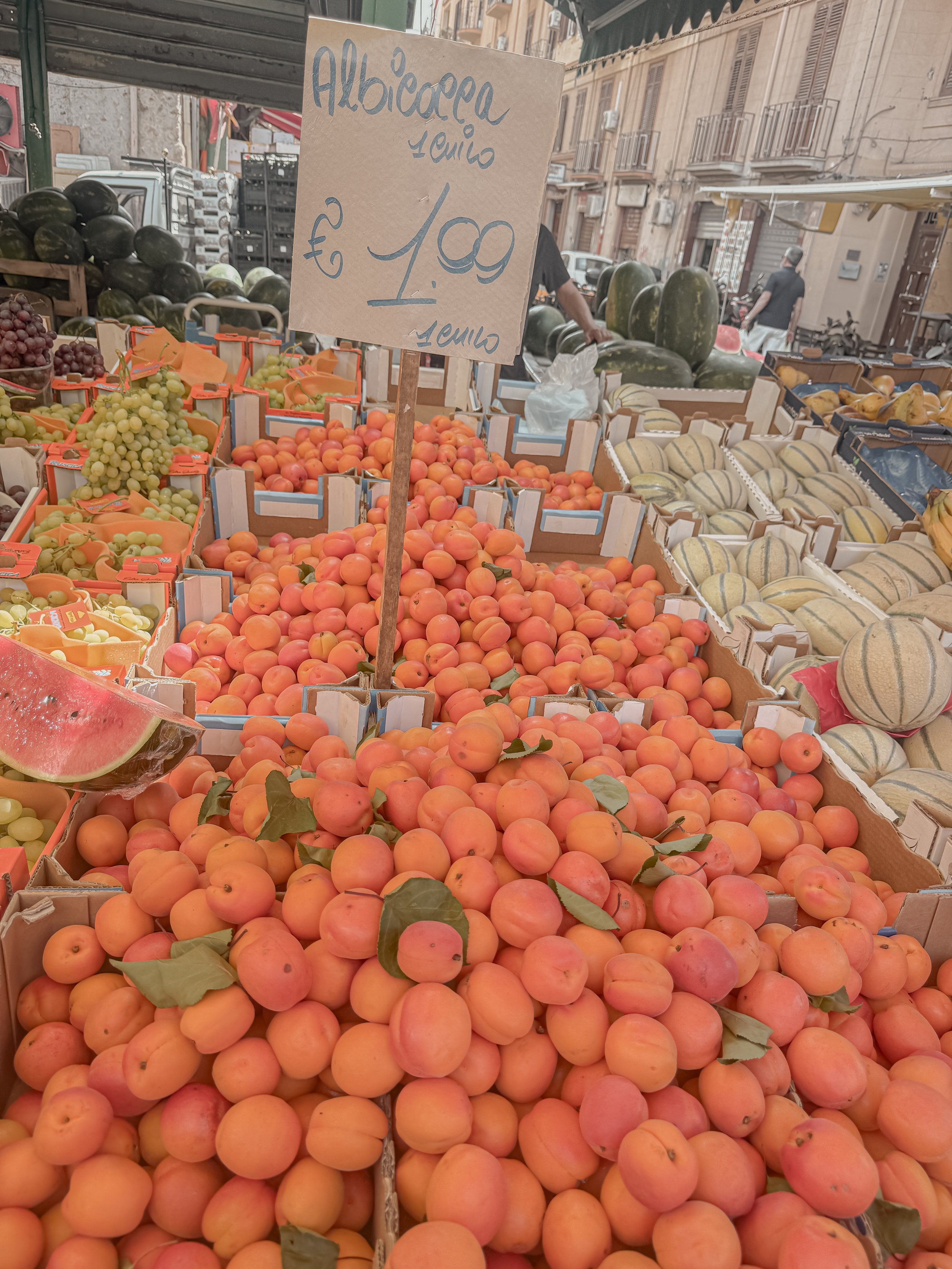 A market stand display of green grapes, purple grapes, watermelons, cantaloupes, and peaches, with a handwritten sign indicating prices per kilogram for apricots in Italian.