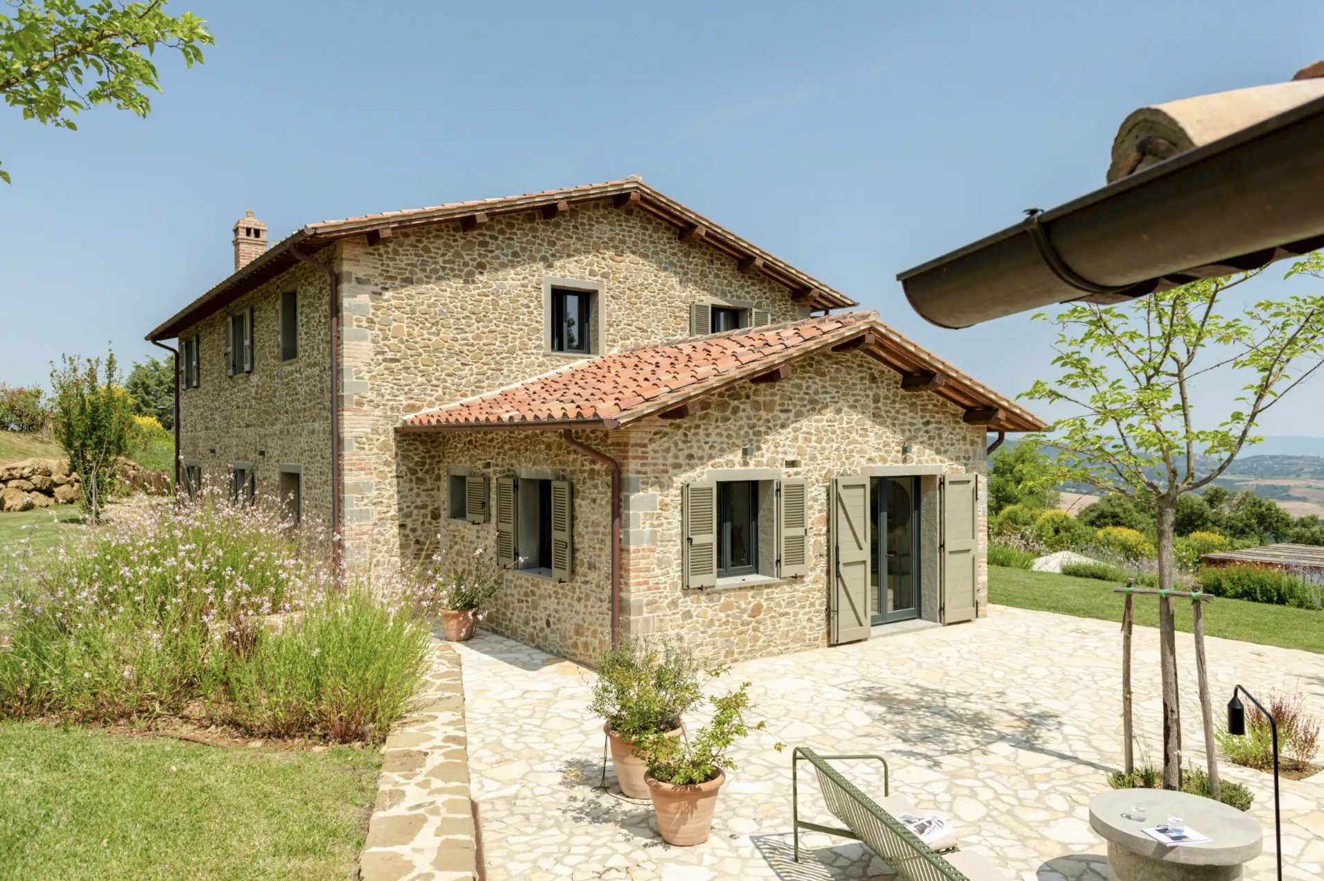 A stone house with a tiled roof, green shutters, and a paved patio surrounded by a garden with plants, trees, and outdoor furniture under a clear blue sky.