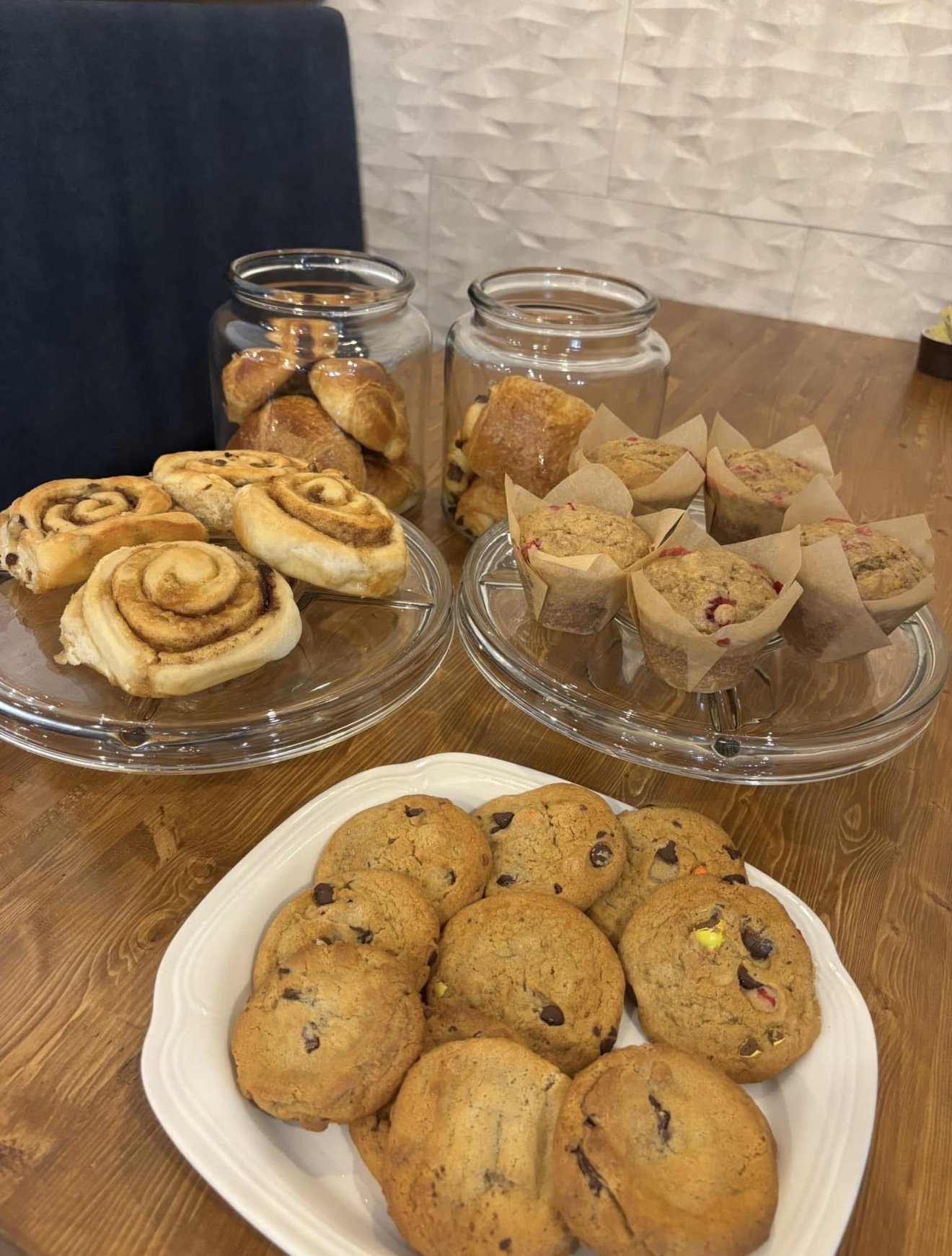 Assortiment de pâtisseries, incluant des roulés à la cannelle, des muffins aux fruits rouges, et des cookies aux pépites de chocolat, sur des plateaux en verre et une assiette en porcelaine sur une table en bois.