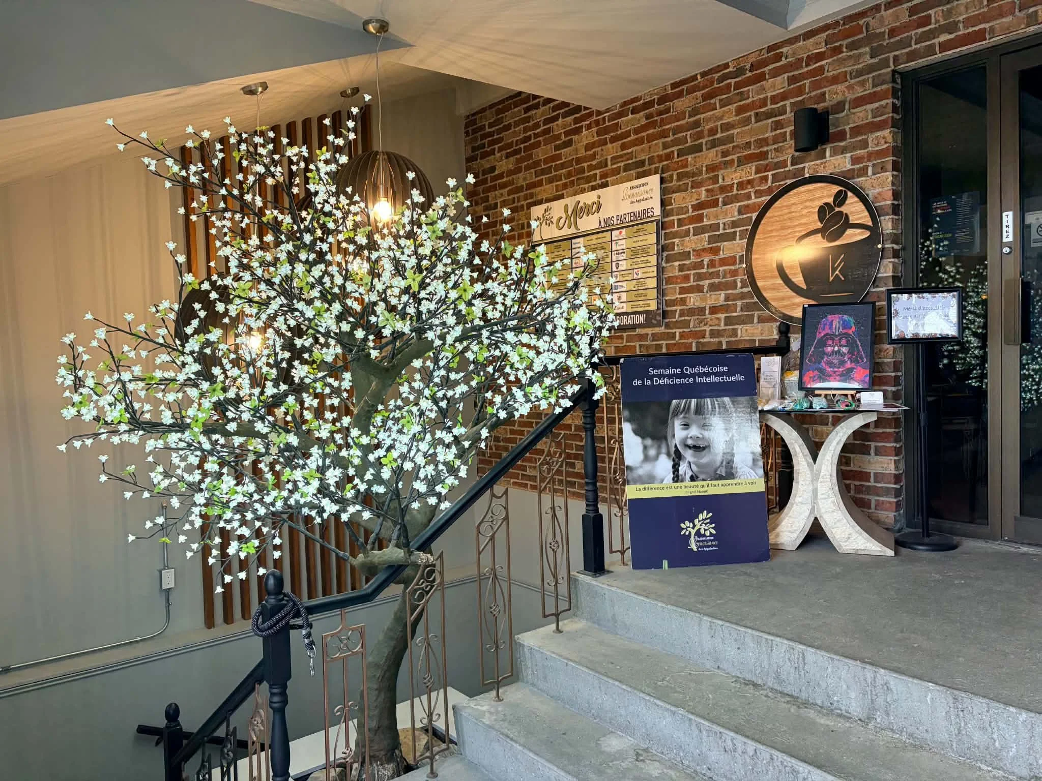 Entrée d'un bâtiment avec un arbre en fleurs artificielles, une affiche de remerciement et des panneaux d'information en français, décorée avec un mur en briques.