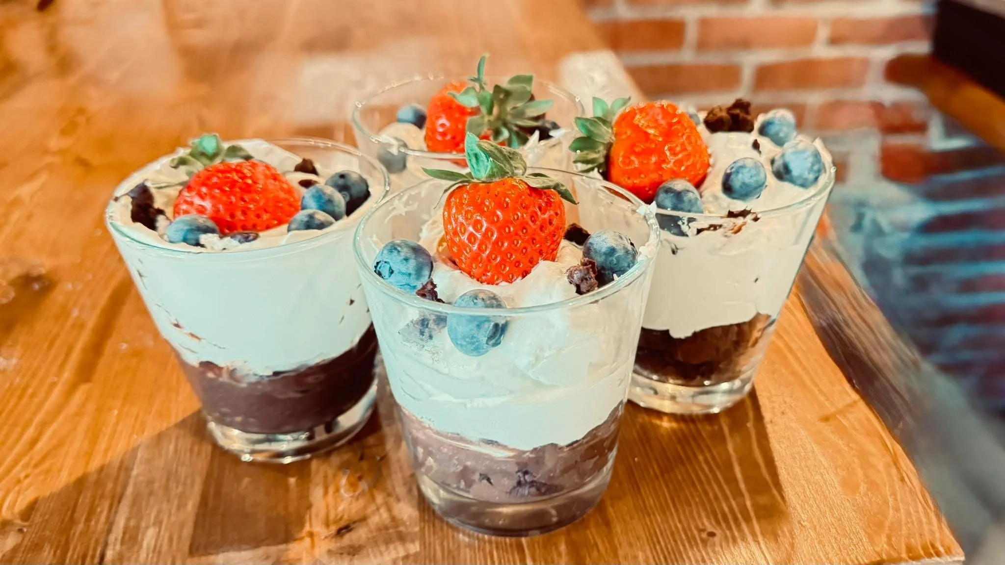 Verres de desserts avec crème, fraises, myrtilles et morceaux de chocolat, sur une table en bois.