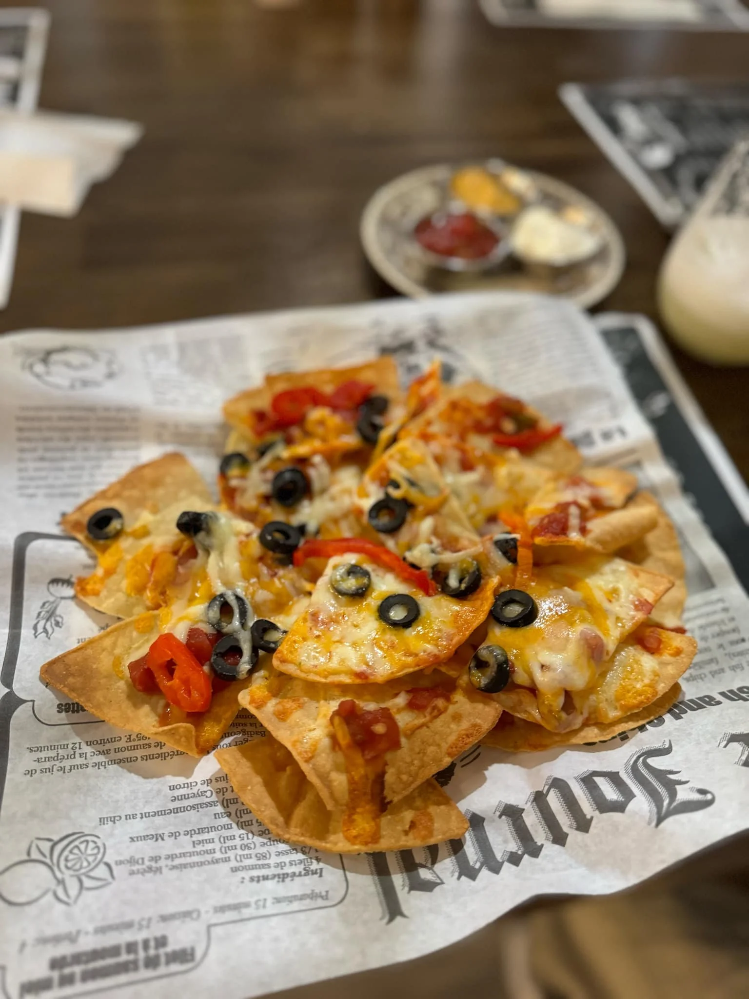 Tacos garnis de fromage, olives noires, poivrons rouges et jambon sur une table en bois avec une assiette de sauces en arrière-plan.