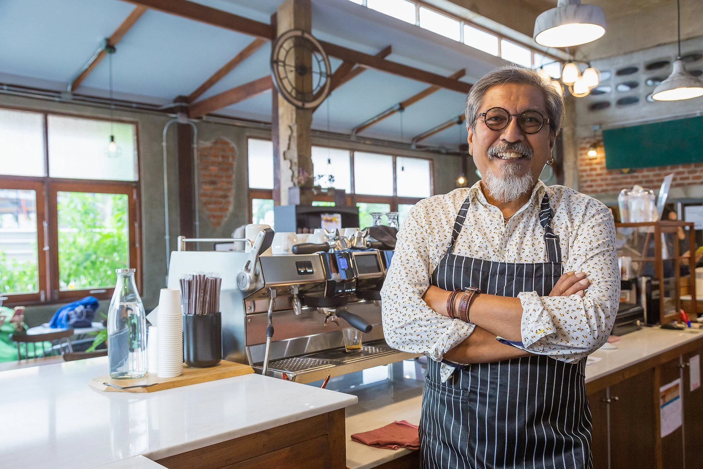 A smiling man with glasses, a white beard, and earrings standing in a cafe with arms crossed, wearing a striped apron over a patterned shirt. The cafe has large windows, exposed brick walls, and industrial-style hanging lights.
