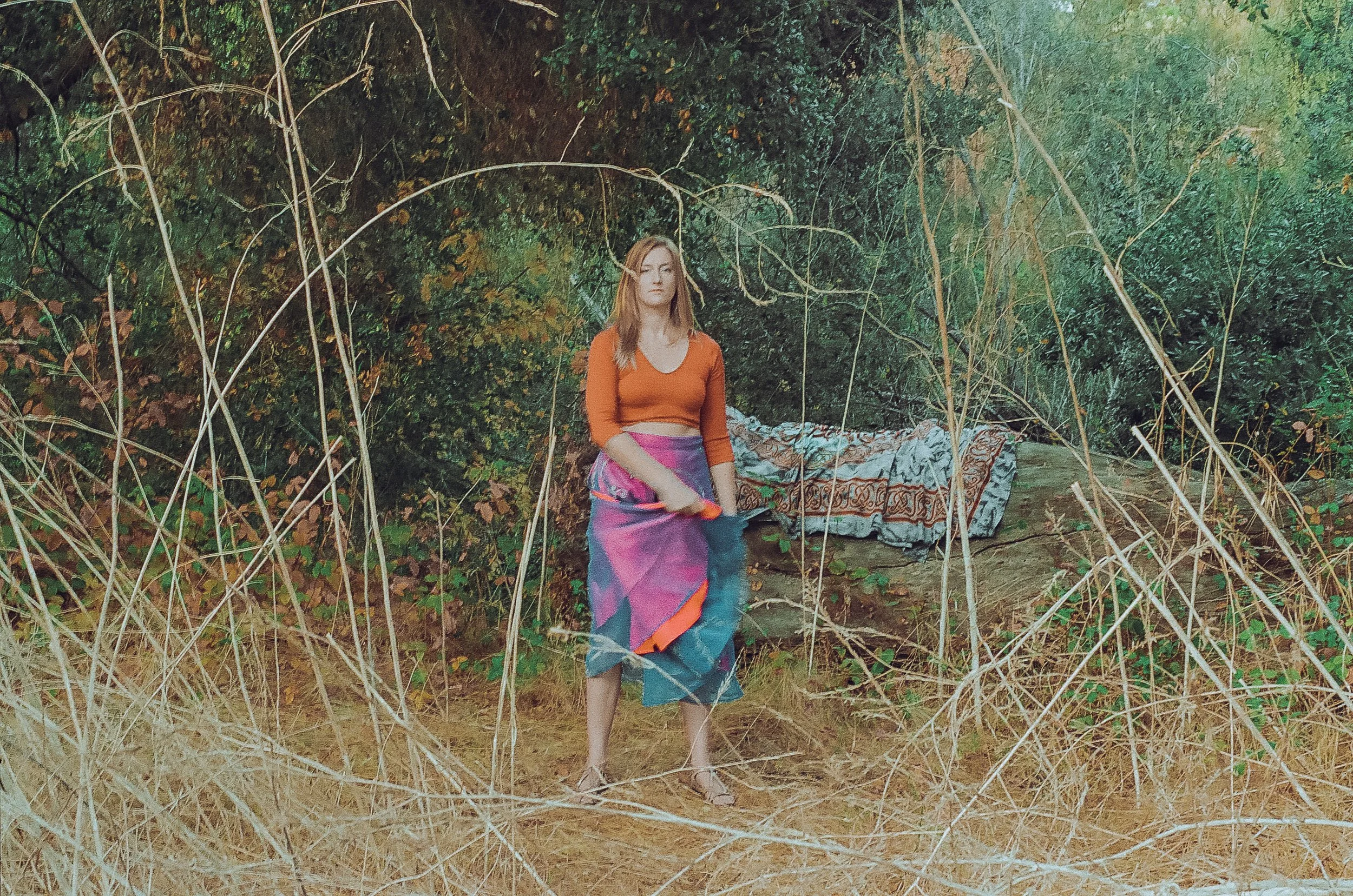 A woman dressed in a colorful skirt and orange top standing outdoors amid tall dry grass and leafy trees.