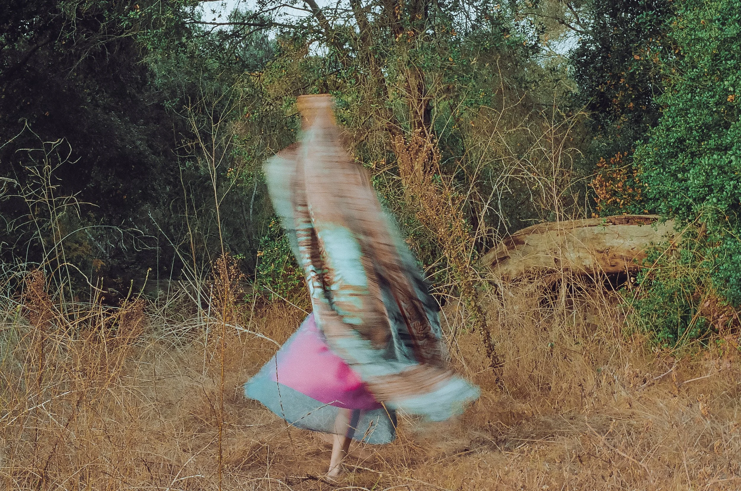 A person wearing a colorful dress is running through a field of dry grass and dense bushes with green leaves.