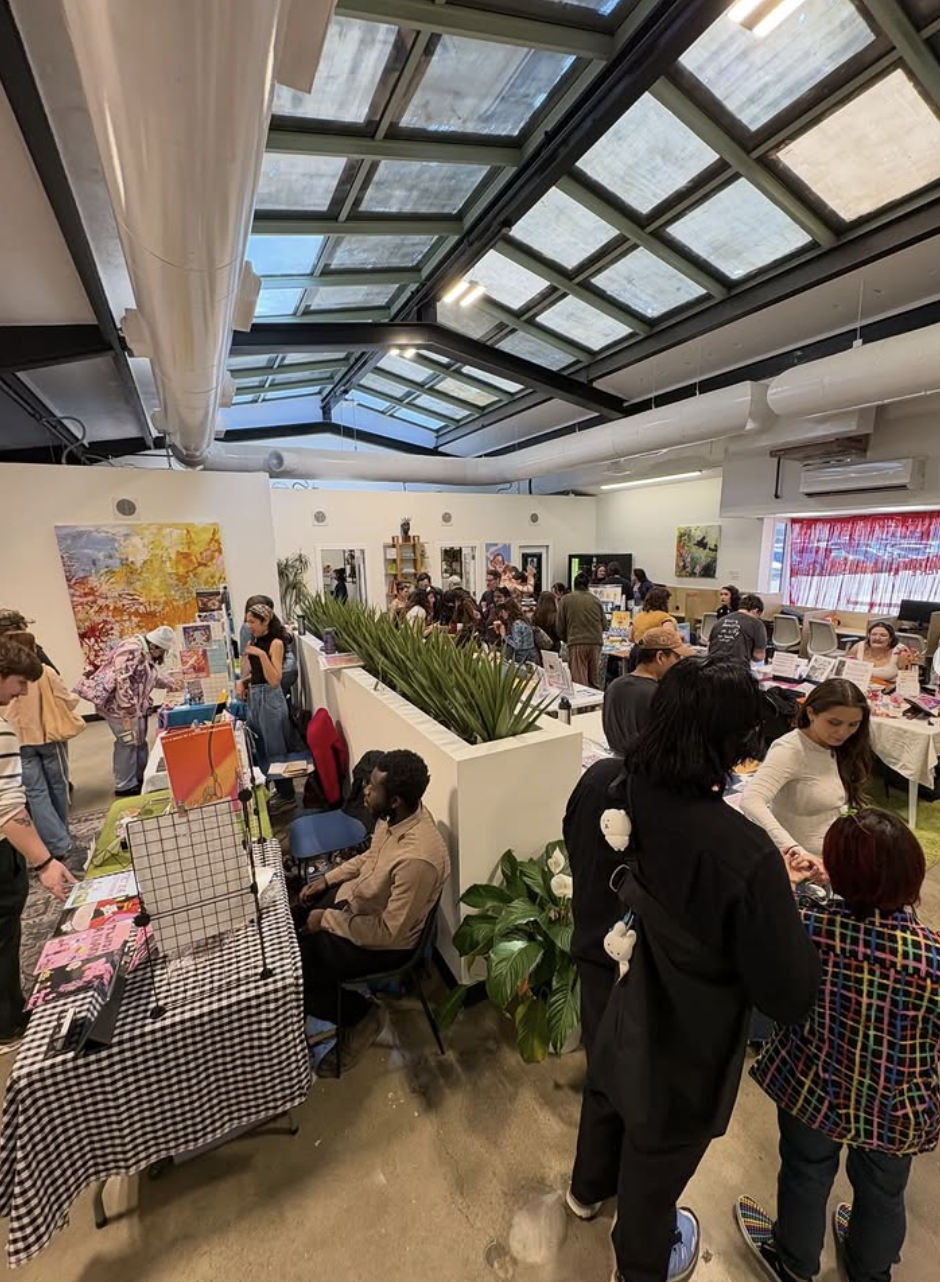 People shopping and browsing at an indoor market or craft fair in a spacious building with a glass ceiling, featuring various booths with art, jewelry, and other items.