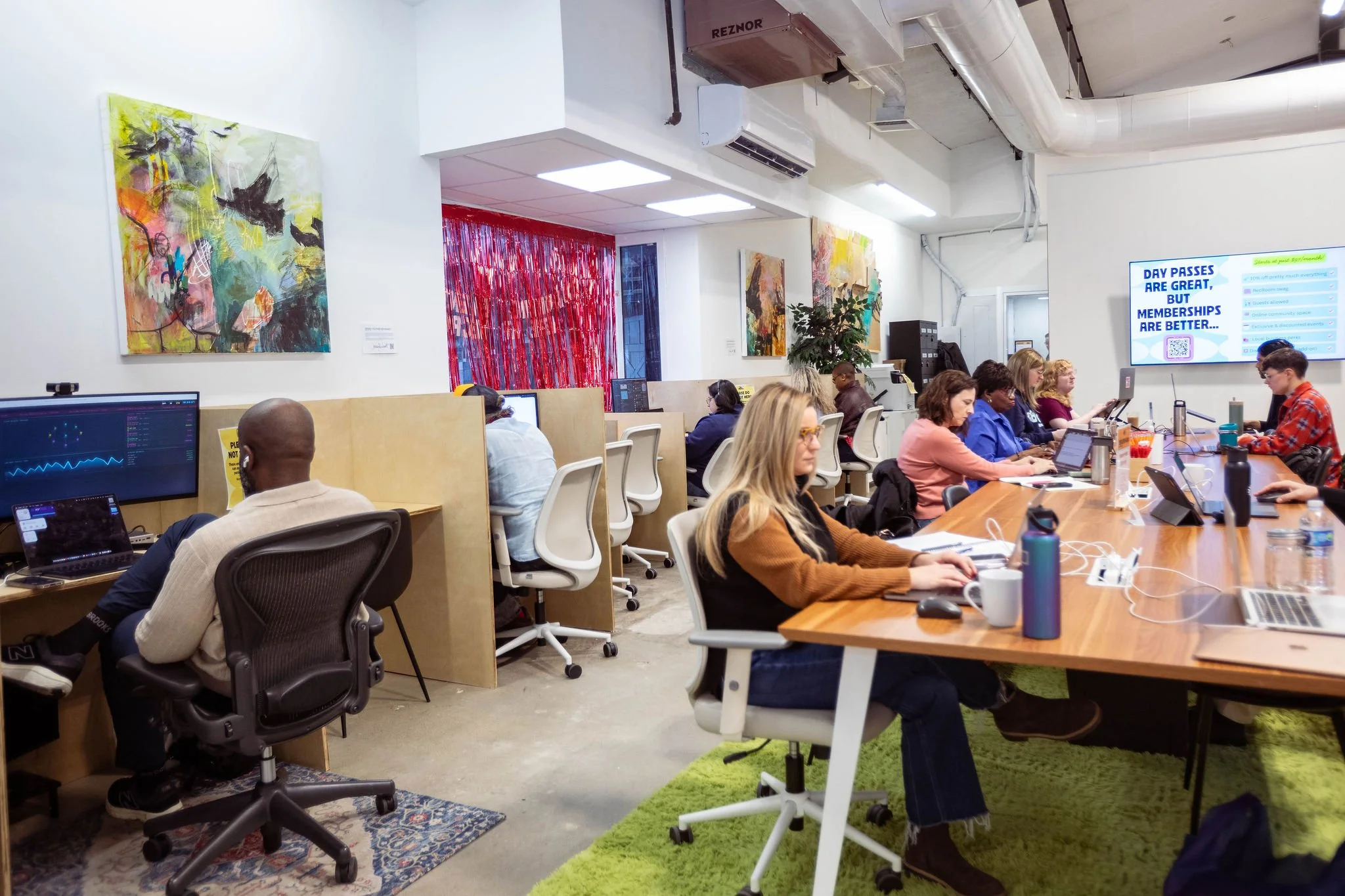 A coworking office space with multiple people working at desks, including computers, with colorful abstract artwork on the walls and an electronic sign displaying a schedule or message.
